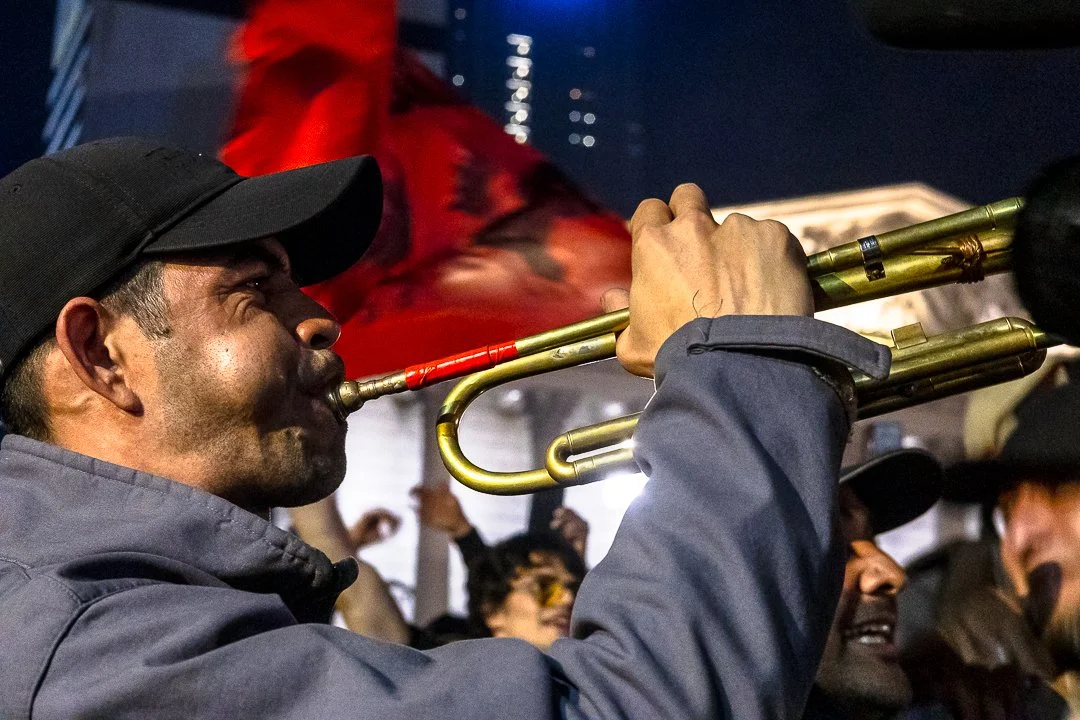 Hombre tocando la trompeta durante una protesta o celebración nocturna, con un grupo de personas en el fondo.