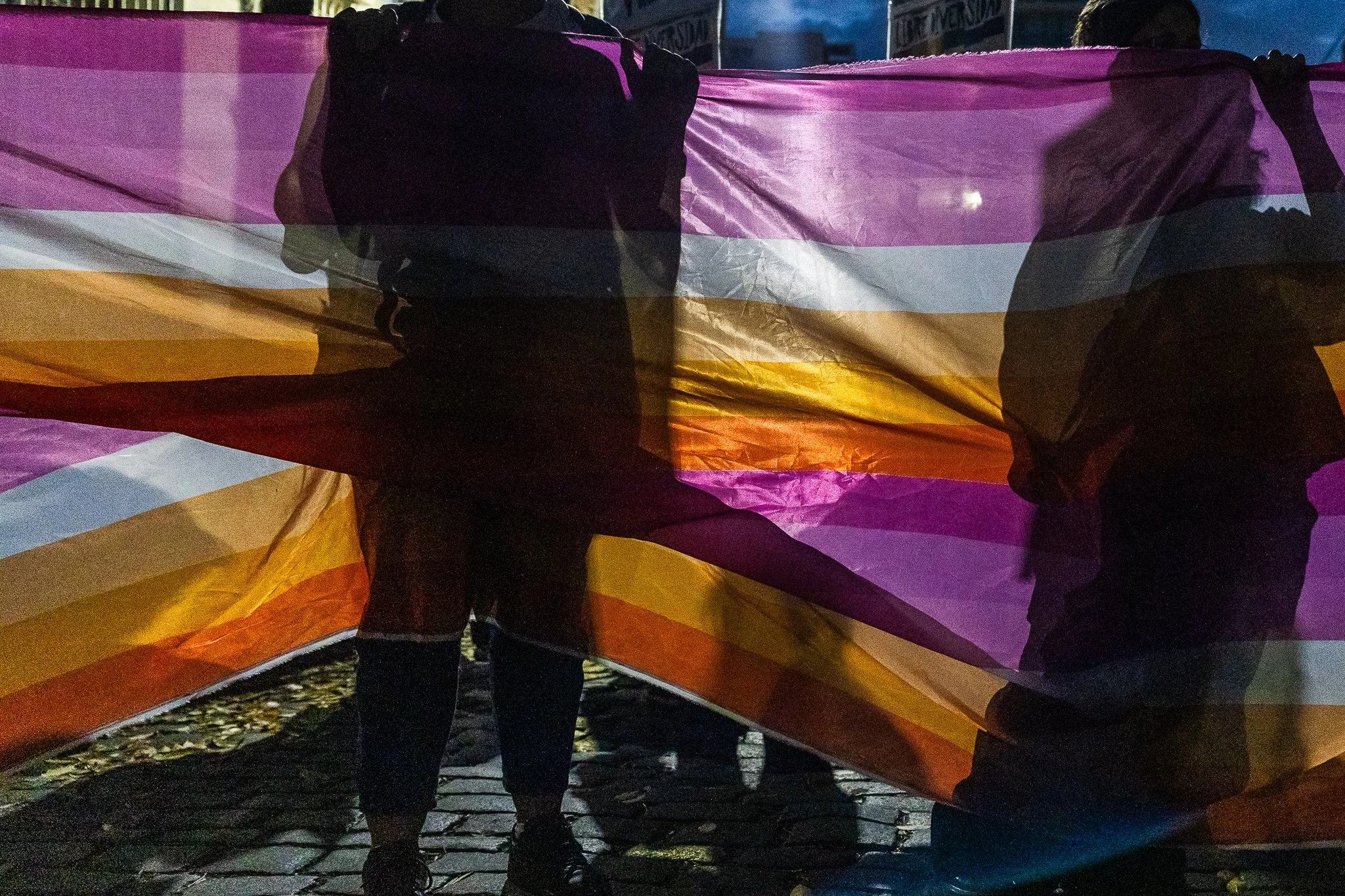 Personas sosteniendo una bandera con los colores de la bandera lesbica: rosa, blanco, fucsia y naranja, en una manifestación en la calle a la noche.