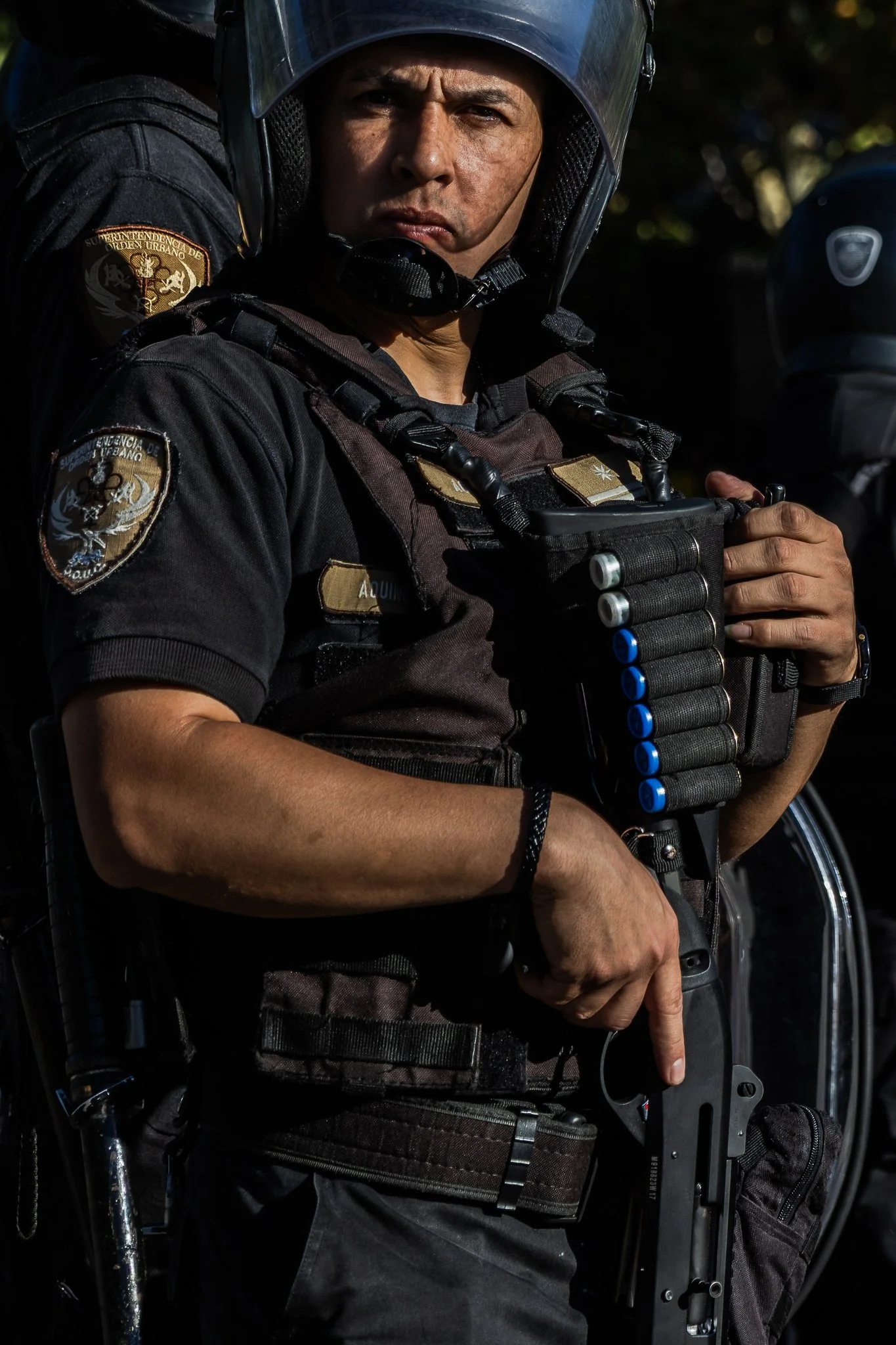 Policía en uniforme con casco y equipo táctico, sosteniendo una escopeta en una postura lista, en un entorno exterior con sombra y luz solar.