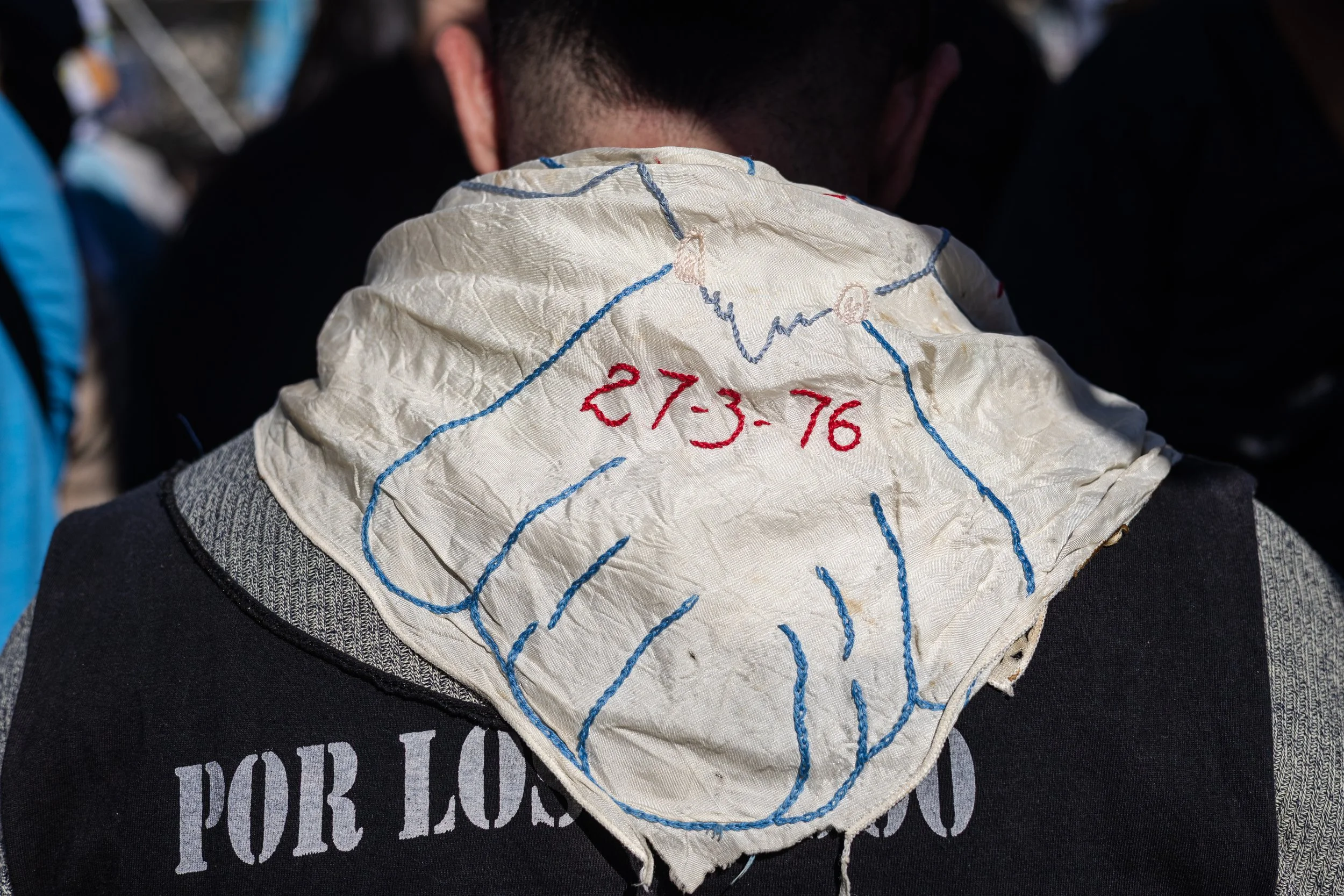 Persona con un pañuelo blanco con bordado azul y rojo en la parte posterior, con la inscripción '27-3-76', viste una sudadera con la leyenda 'POR LOS QUE'.