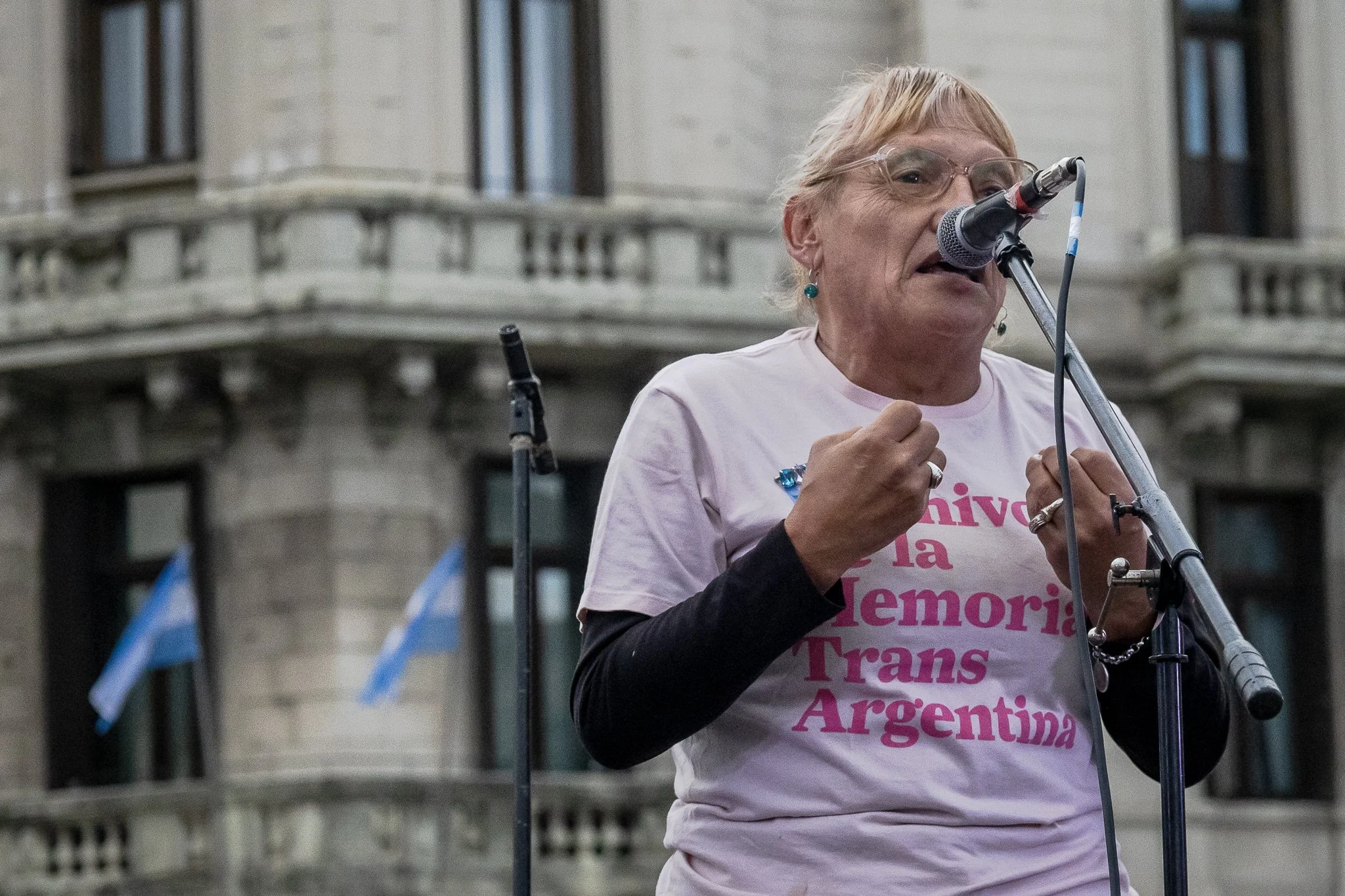 Mujer con gafas y ropa clara participando en una manifestación o concentración en la calle, sosteniendo un megáfono, con un edificio de fondo y banderas argentinas.