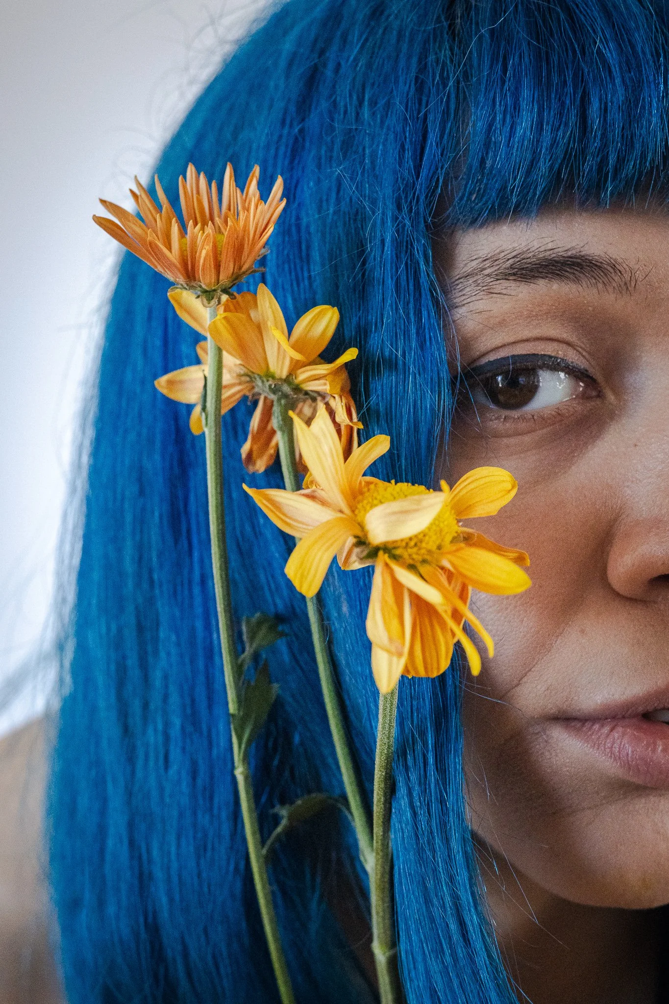Una persona con cabello azul brillante, parcialmente visible, tiene flores amarillas y anaranjadas colocadas cerca de su rostro.