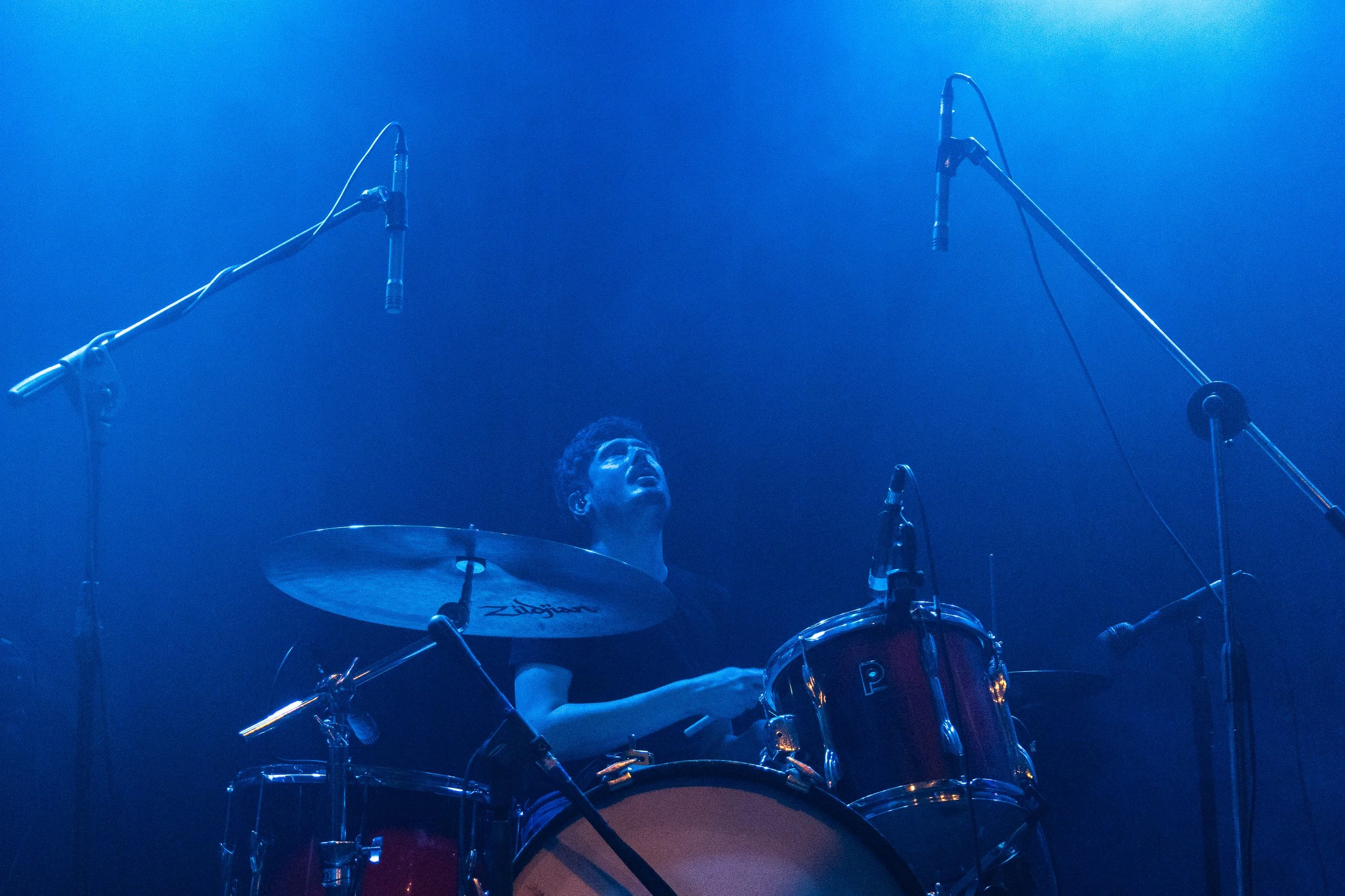 Baterista tocando en un escenario con iluminación azul, rodeado de micrófonos