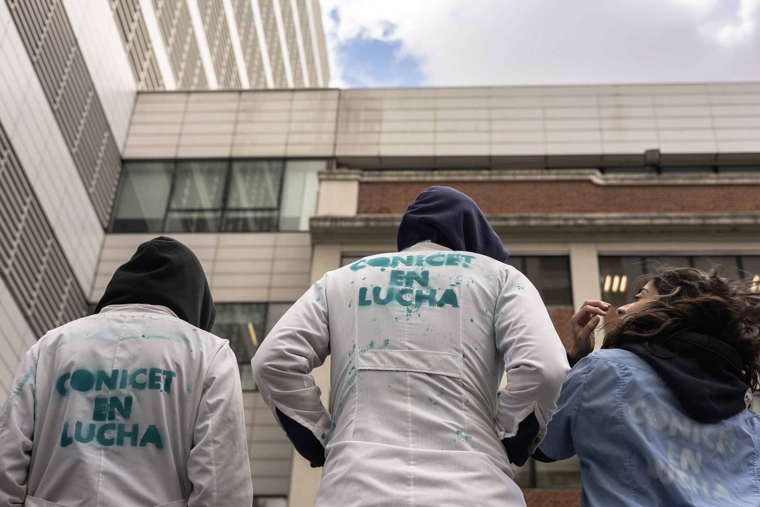 Tres personas con ropa blanca y una con sudadera azul, de espaldas, en una protesta en un edificio moderno, una con cabello largo y rizado.