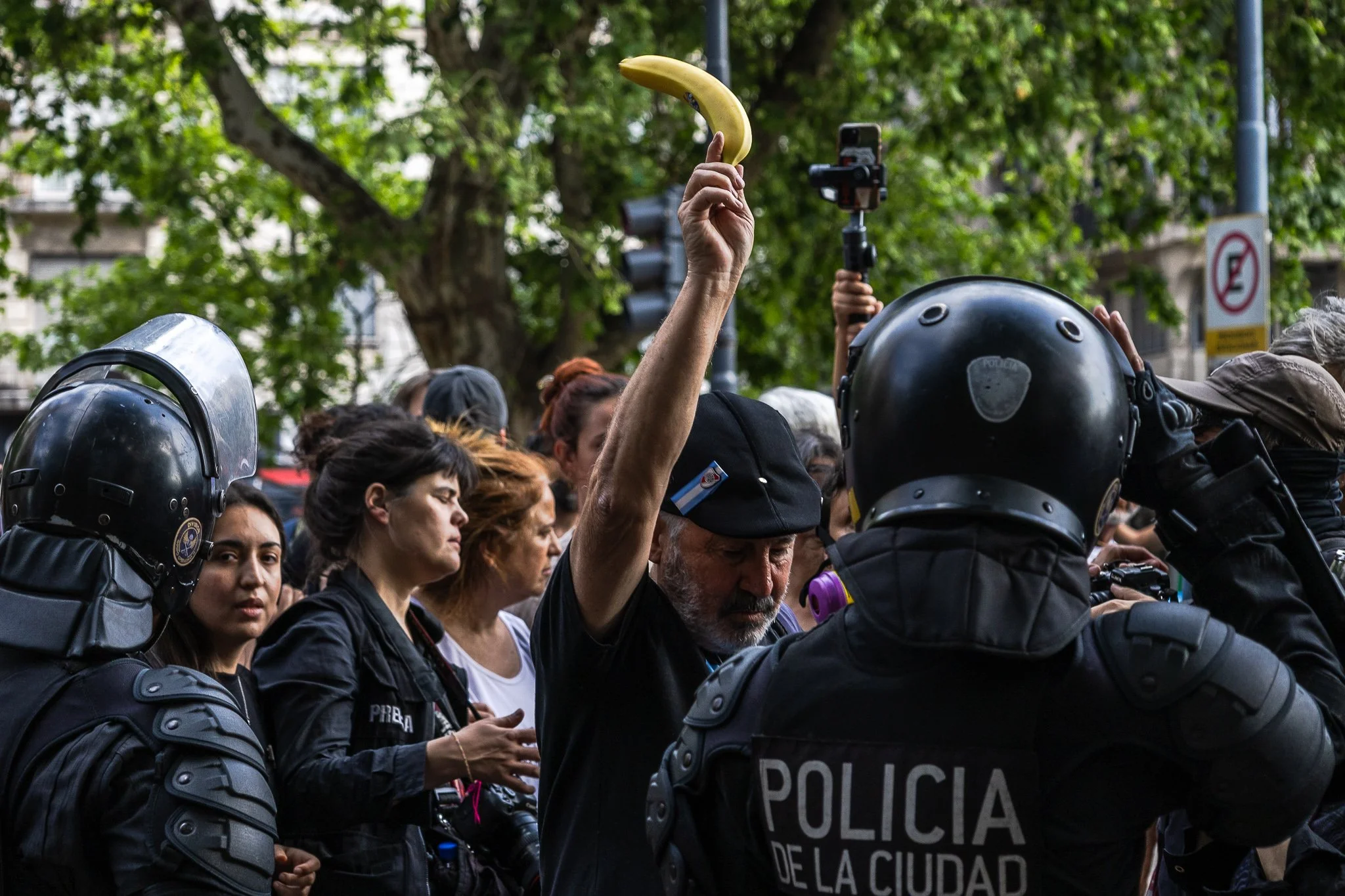 Manifestantes pacíficos enfrentándose a la policía en un espacio público con árboles y señales de tráfico en el fondo.