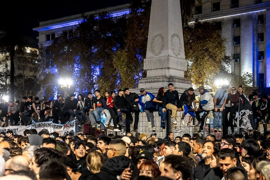 Muchos jóvenes protestando en la calle, algunos se encuentran en una valla de seguridad y otros en el suelo. Hay árboles y edificios iluminados en el fondo, de noche.