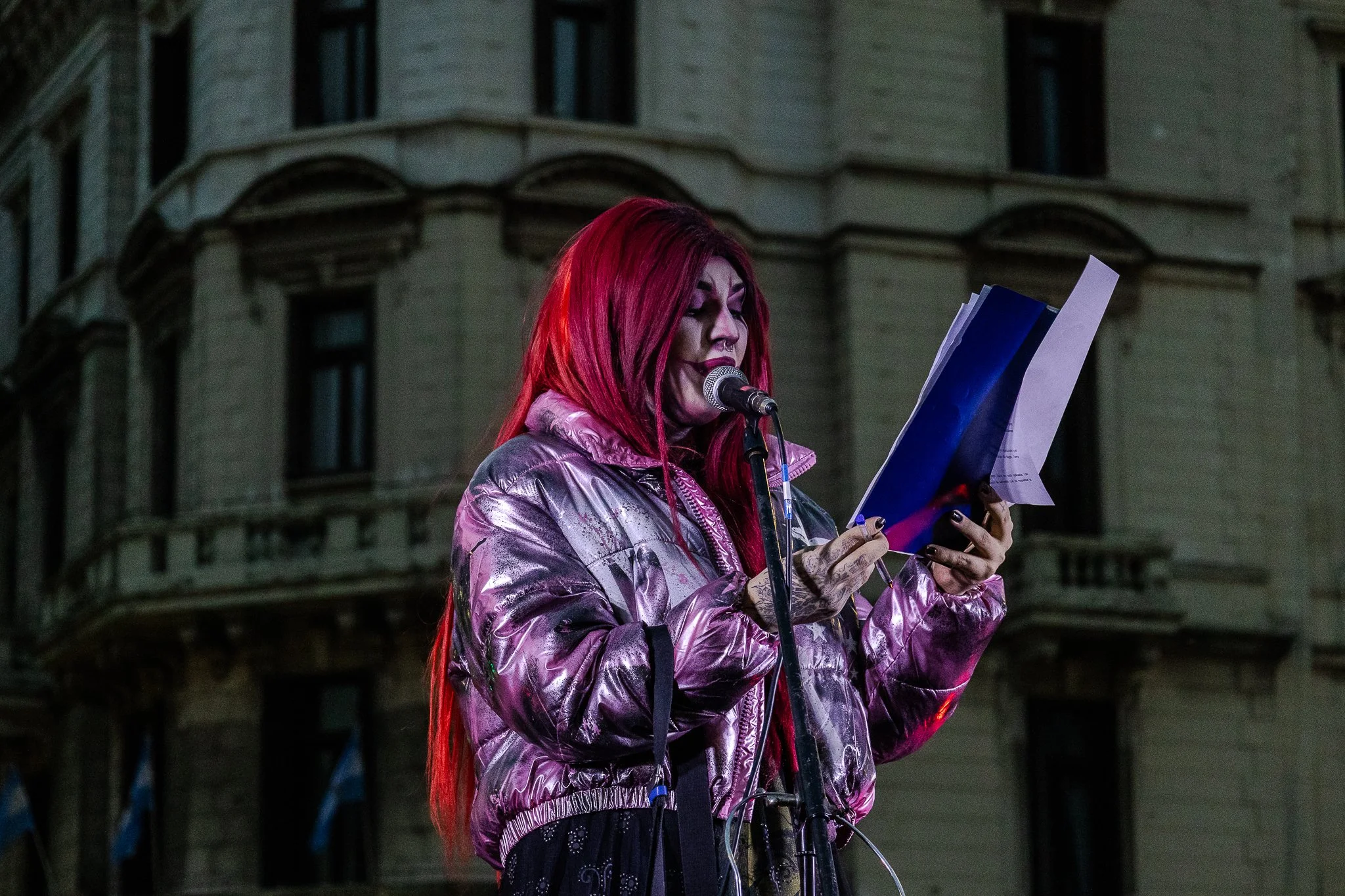 Mujer con cabello largo y teñido de rojo, vestida con abrigo plateado, leyendo un papel frente a un micrófono en la calle, con un edificio antiguo de fondo.