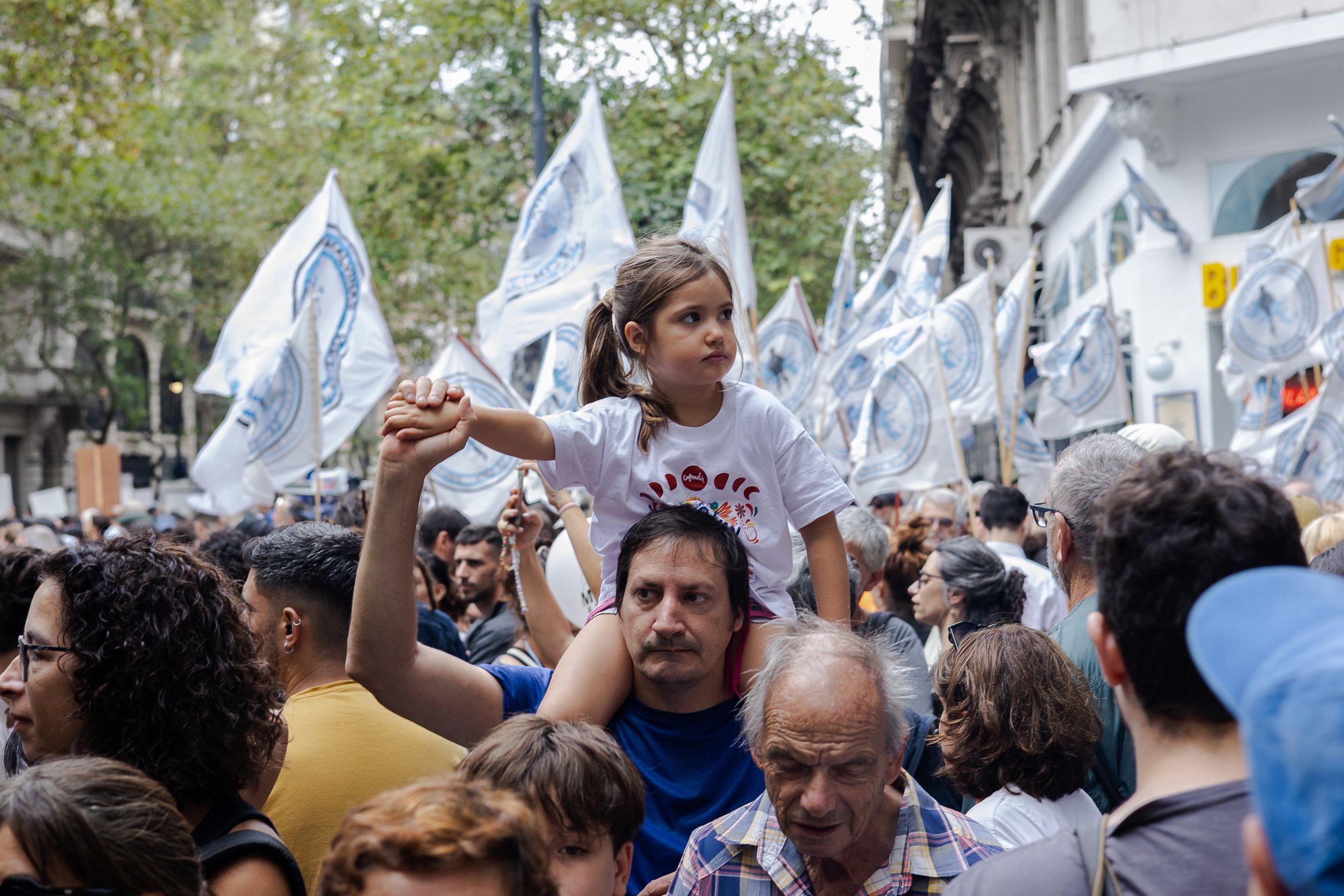 Una multitud en una manifestación con muchas banderas blancas, una niña sentada en los hombros de un hombre en primer plano, rodeados de personas diversas.