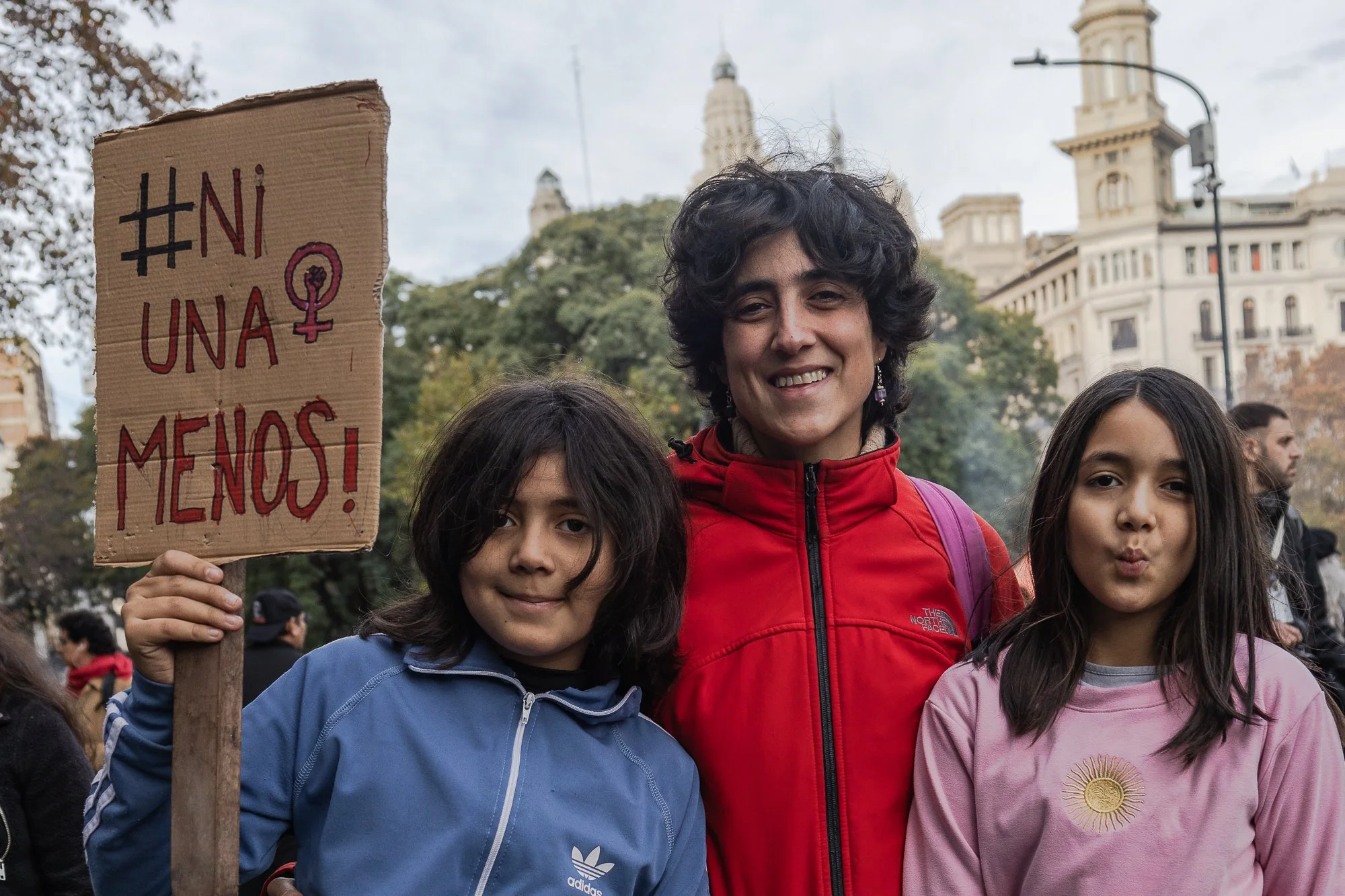 Grupo de personas participando en una manifestación o protesta en la calle. Una persona sostiene un cartel con el hashtag #NiUnaMenos. La escena tiene árboles y edificios históricos en el fondo.