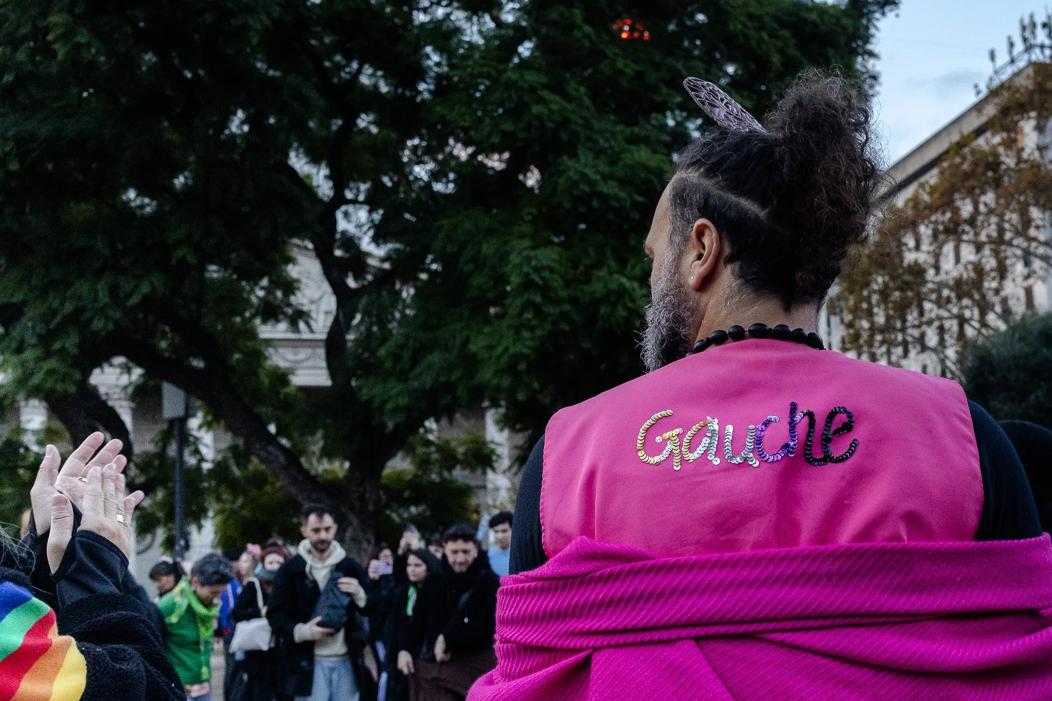 Persona con peinado de cabello rizado y barba, vistiendo un chaleco rosa con la palabra "Gauche" en lentejuelas, entre un grupo de personas en un entorno al aire libre.