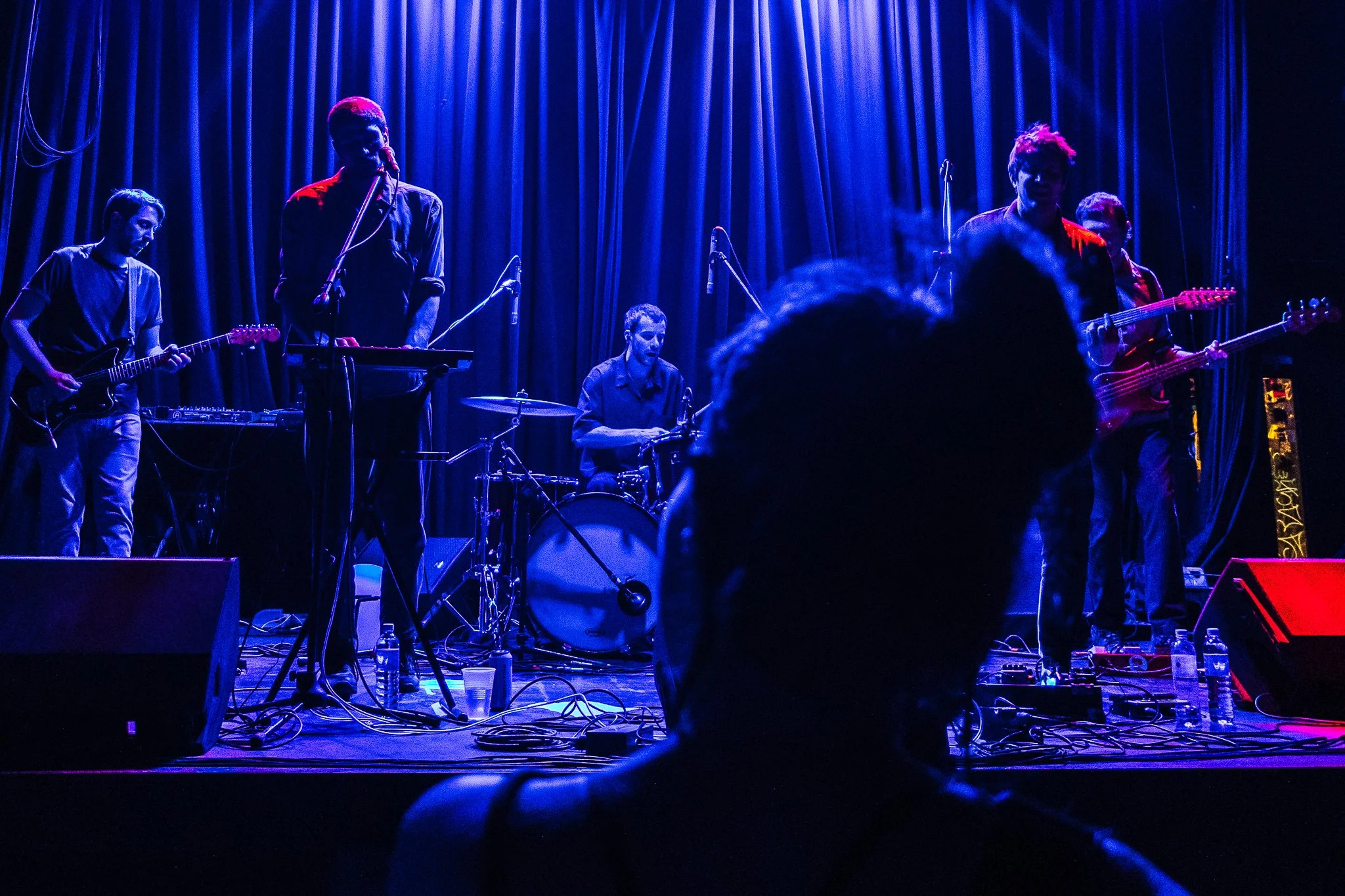 Un grupo de músicos en un escenario con cortinas azules, tocando instrumentos en un concierto.