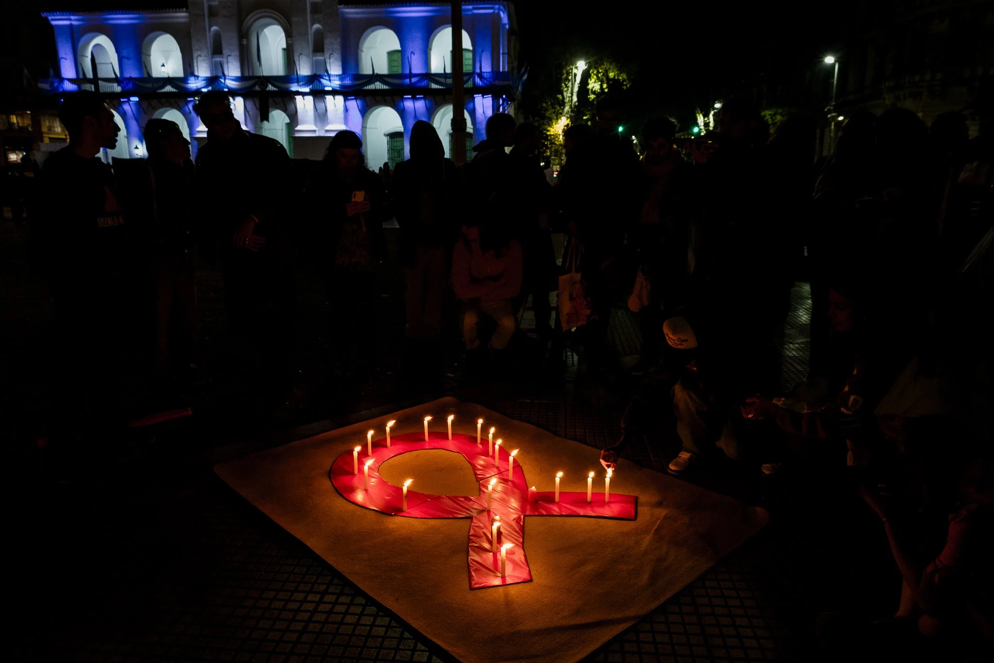 Grupo de personas en la noche en una calle con luces y un edificio iluminado al fondo. En el suelo, hay una señal de tránsito con velas encendidas formando un corazón y un símbolo de paz.
