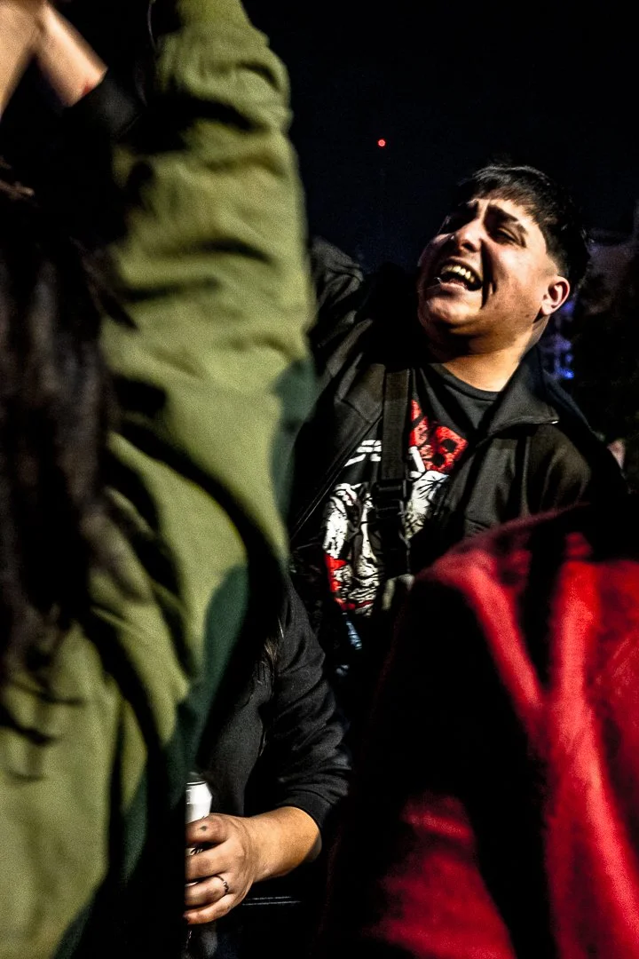 Joven con expresión emocional en un entorno nocturno, rodeado de otras personas con ropa de colores oscuros y rojos.