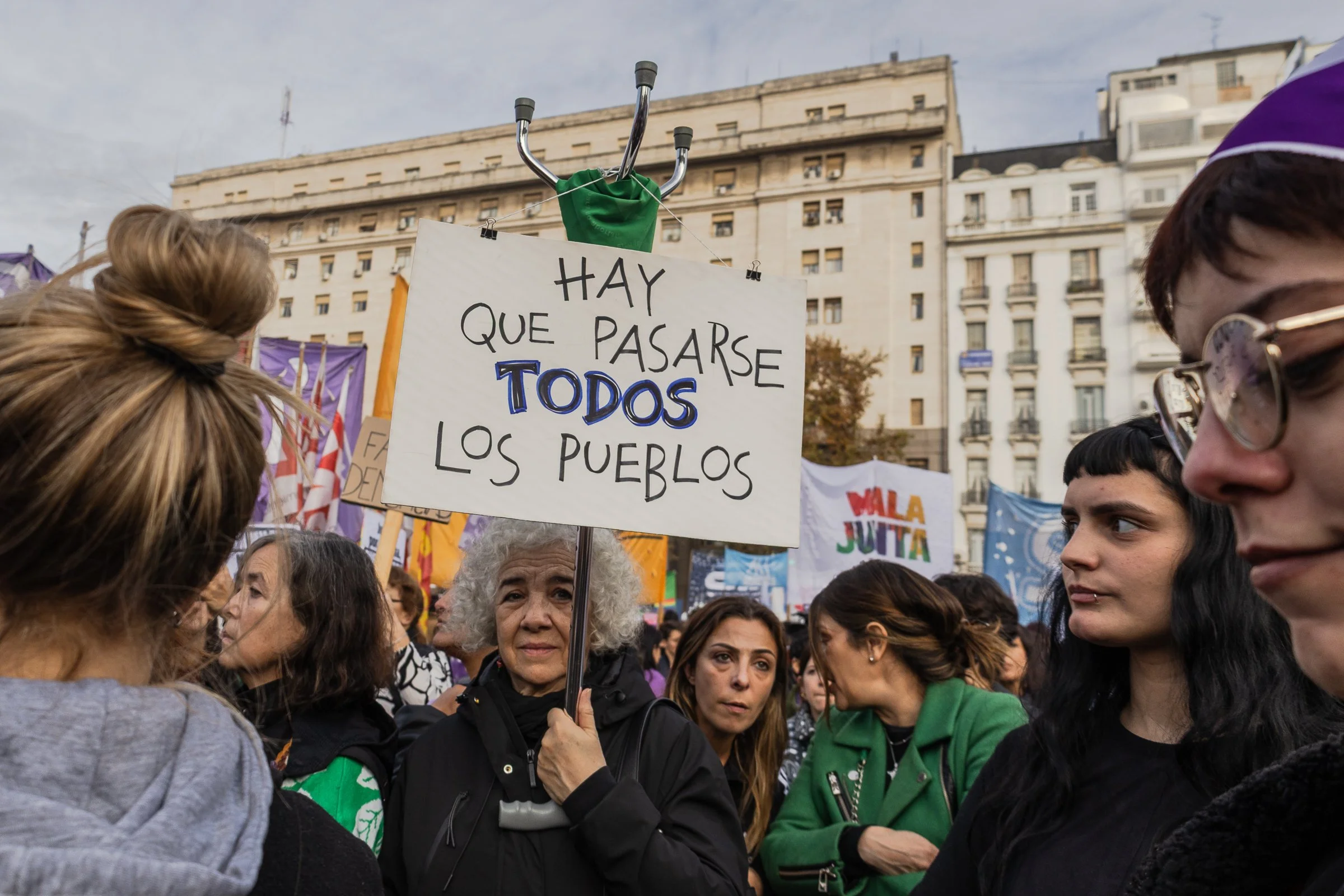 Una mujer mayor sostiene un cartel que dice 'Hay que pasarse todos los pueblos'. Hay varias personas alrededor participando en una marcha o protesta en una plaza urbana con edificios de fondo.