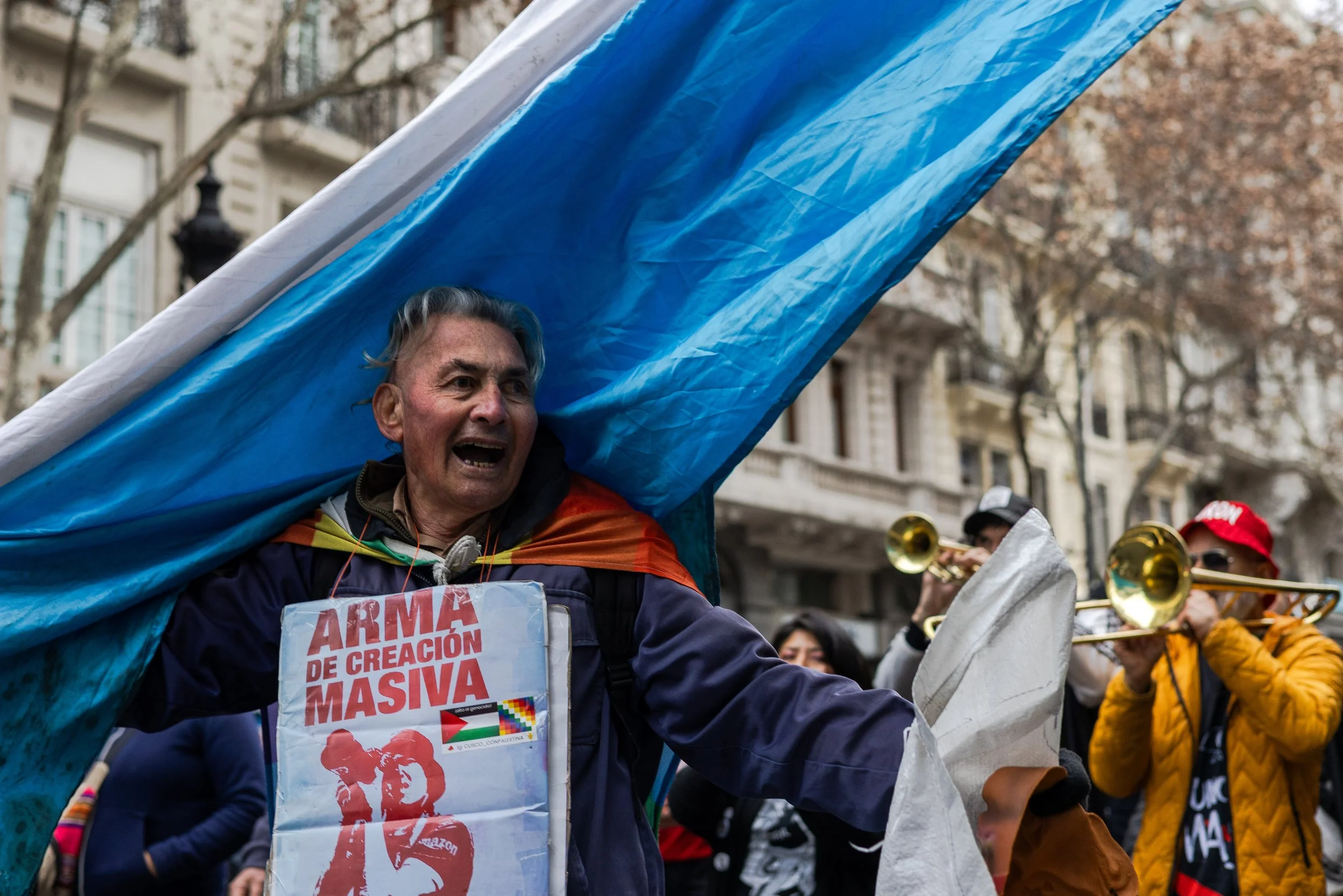 Persona participando en manifestación, llevando una pancarta que dice 'ARMAS DE CREACIÓN MASIVA', durante una marcha en la calle, con árboles y edificios al fondo.