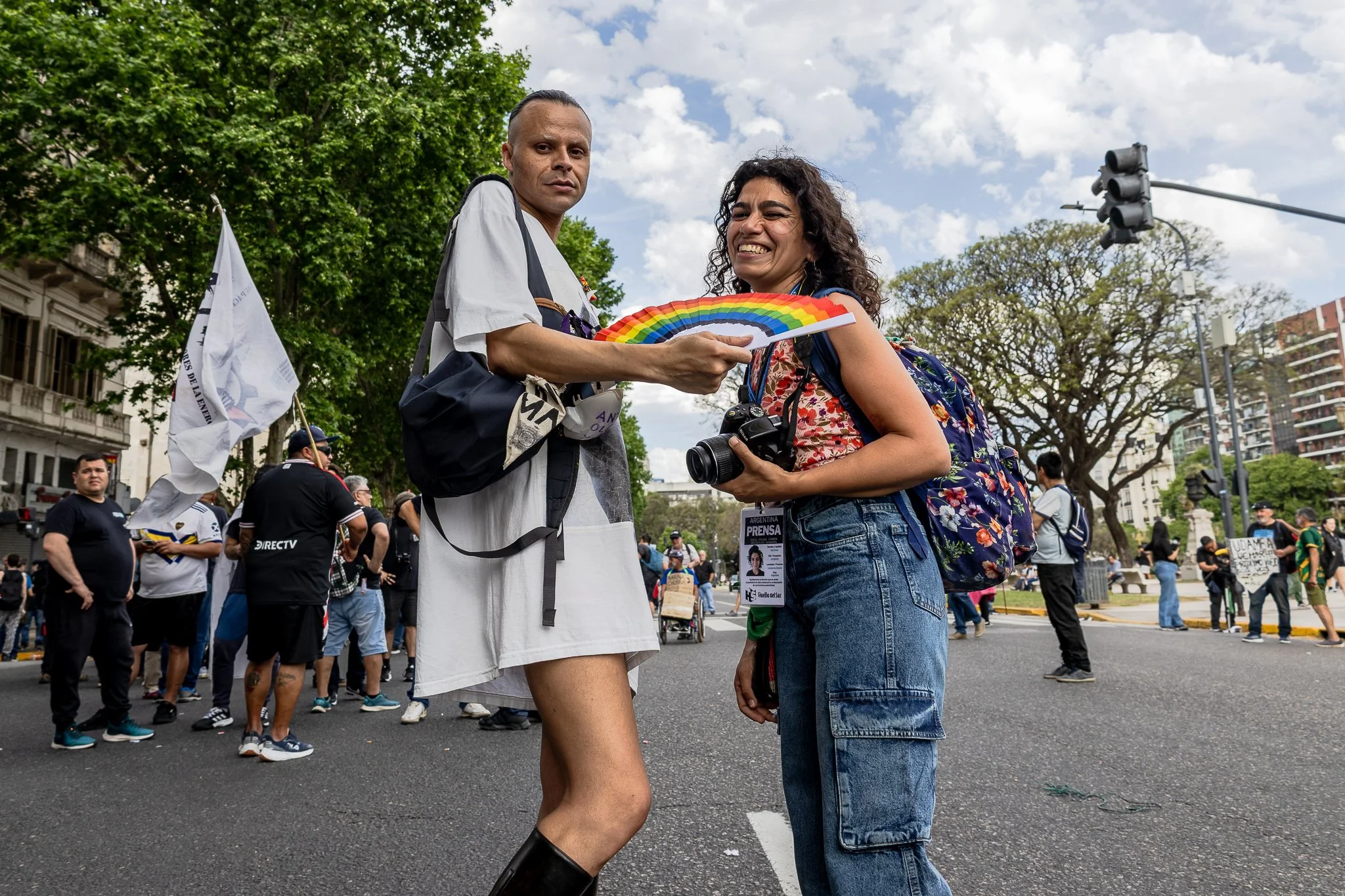 Dos personas en una calle participando en una manifestación, una de ellas sostiene un arcoíris de papel y la otra una cámara, en medio de un grupo de manifestantes.