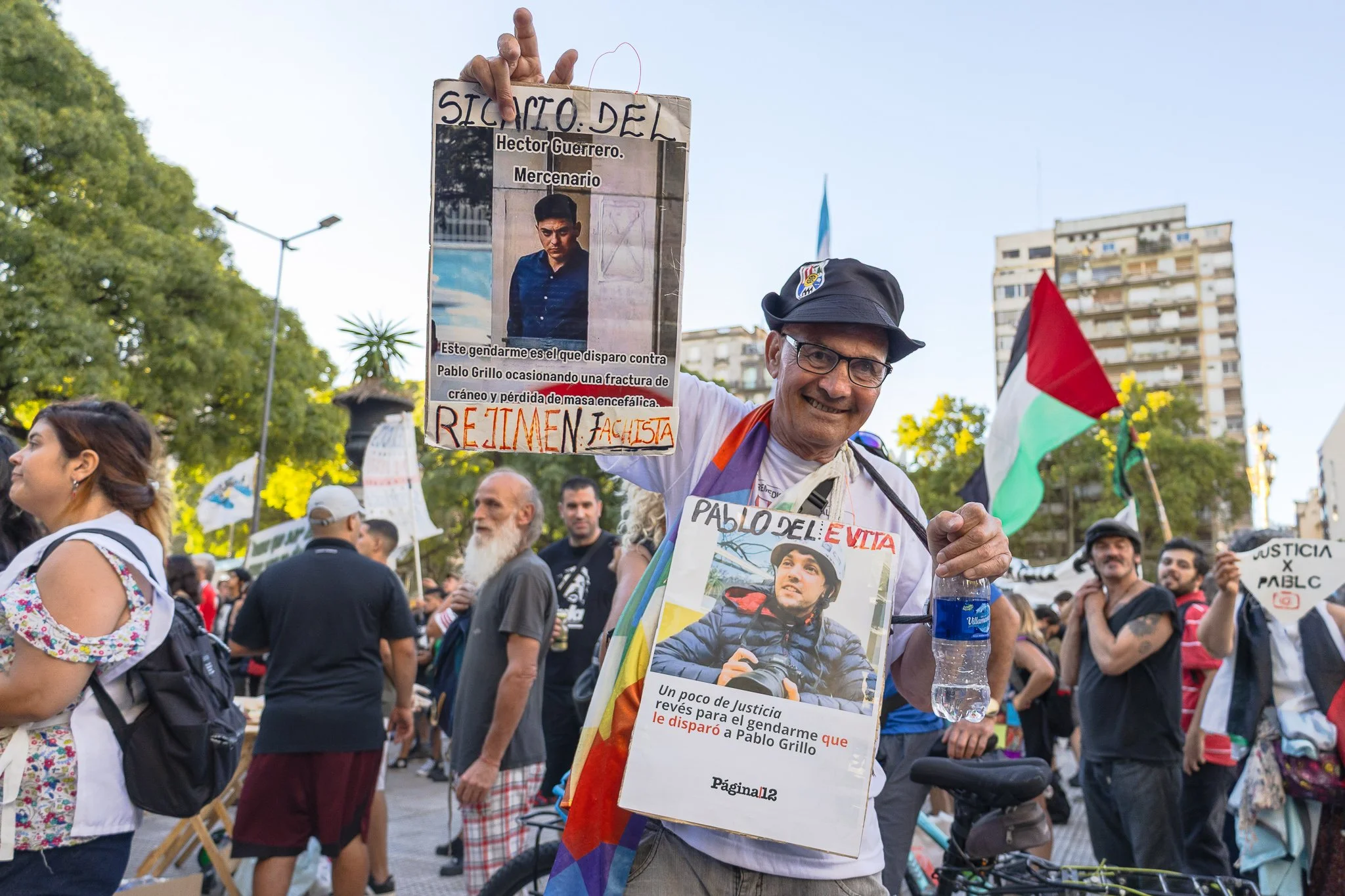 Hombre con gafas, sombrero y camiseta blanca sostiene cartel de protesta que tiene fotos y texto, entre una multitud en una plaza, con edificios y árboles en el fondo.