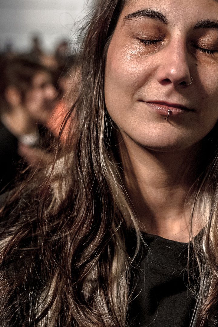 Retrato de una mujer con ojos cerrados y llorando, cabello largo y rizado, con piercings en la nariz y en el labio, en un ambiente difuso y con otras personas al fondo.