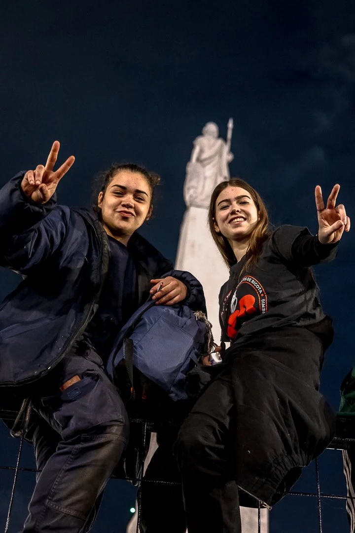 Dos jóvenes en ropa de abrigo posando con victoria y haciendo señal de paz, en un lugar con estatua y cielo nocturno.