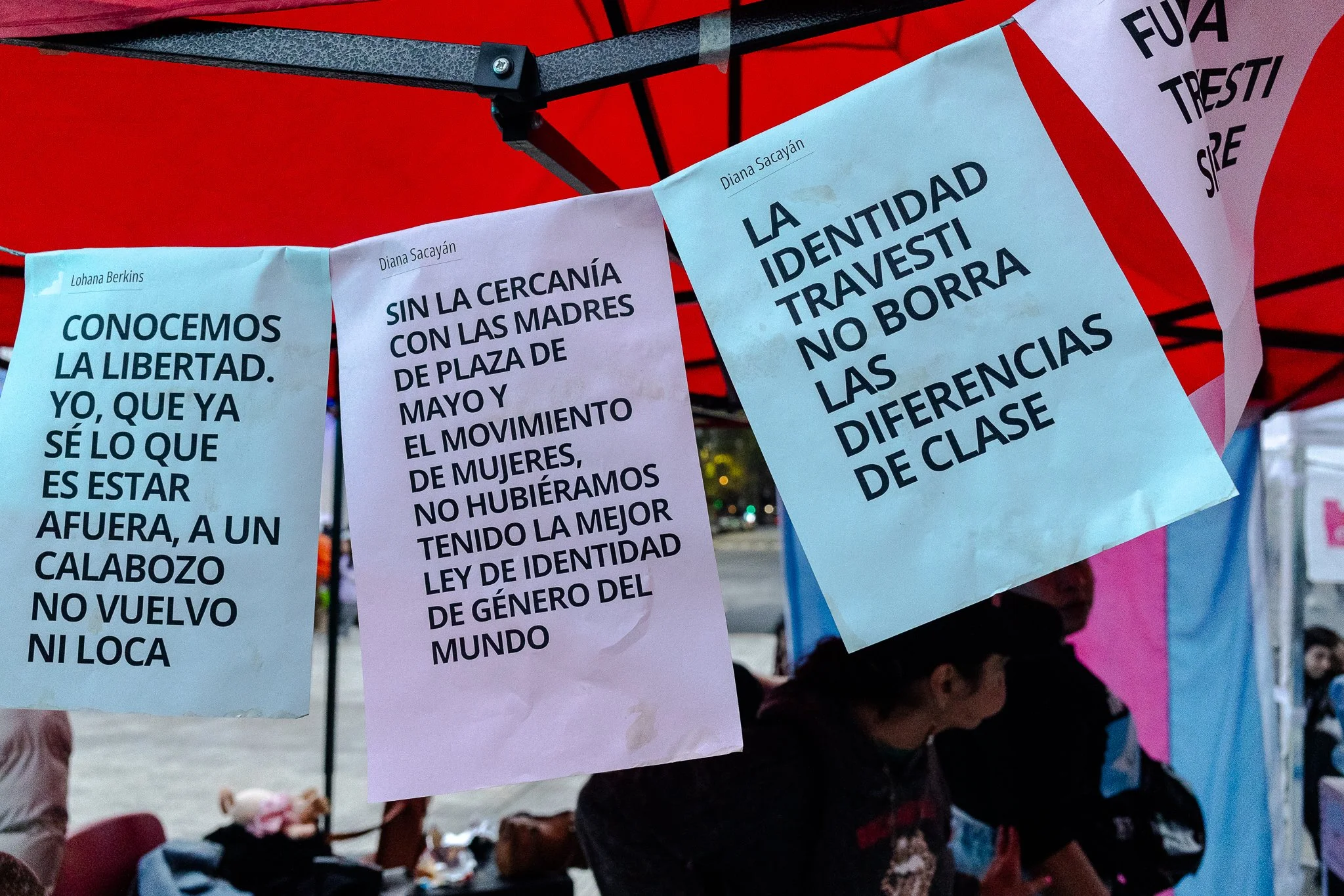 Olas con varios carteles de papel colgados, con mensajes en español sobre libertad, identidad y derechos de las mujeres trans y travestis, en un evento al aire libre con varias personas.