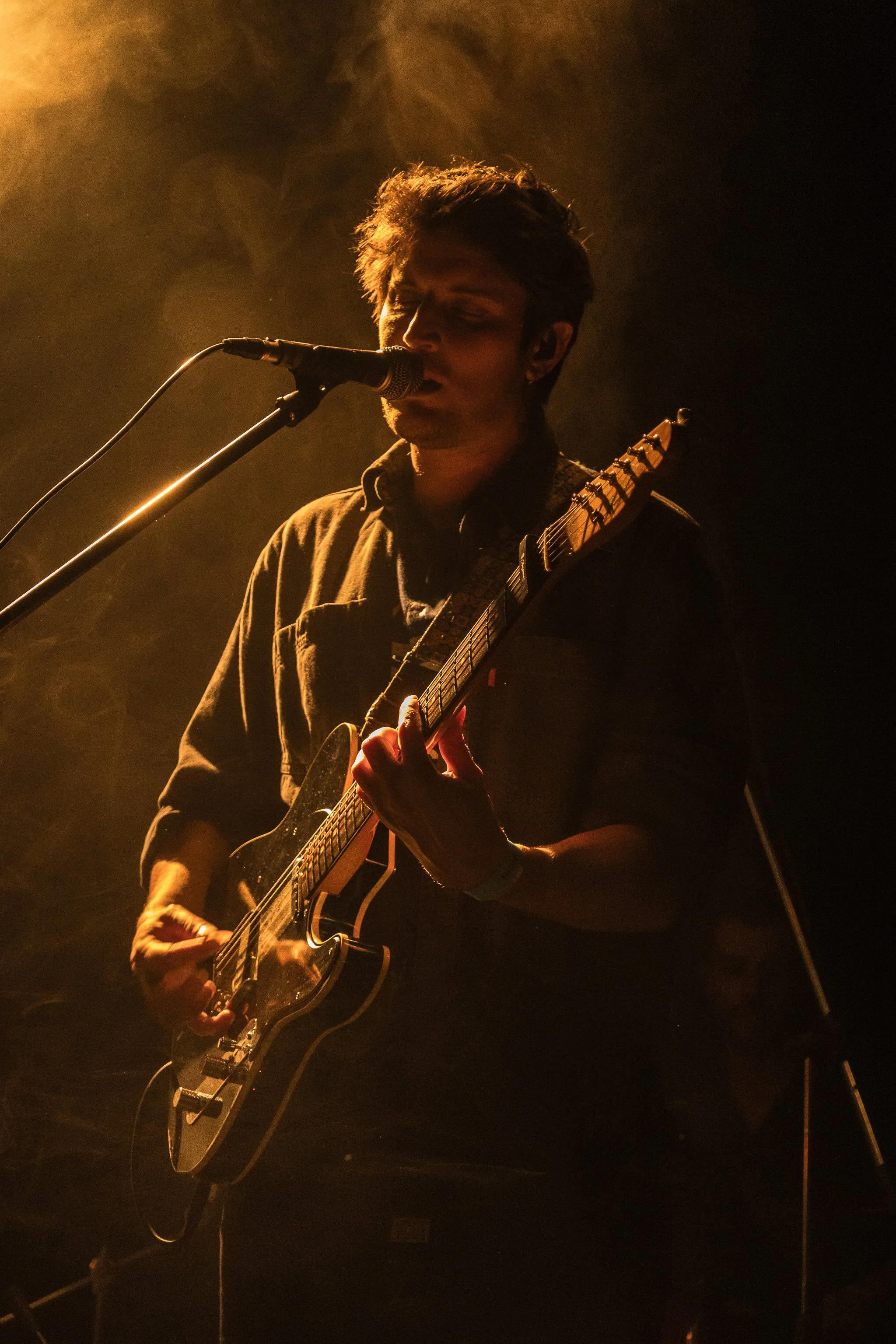 Joven tocando guitarra eléctrica y cantando en un escenario con iluminación cálida.