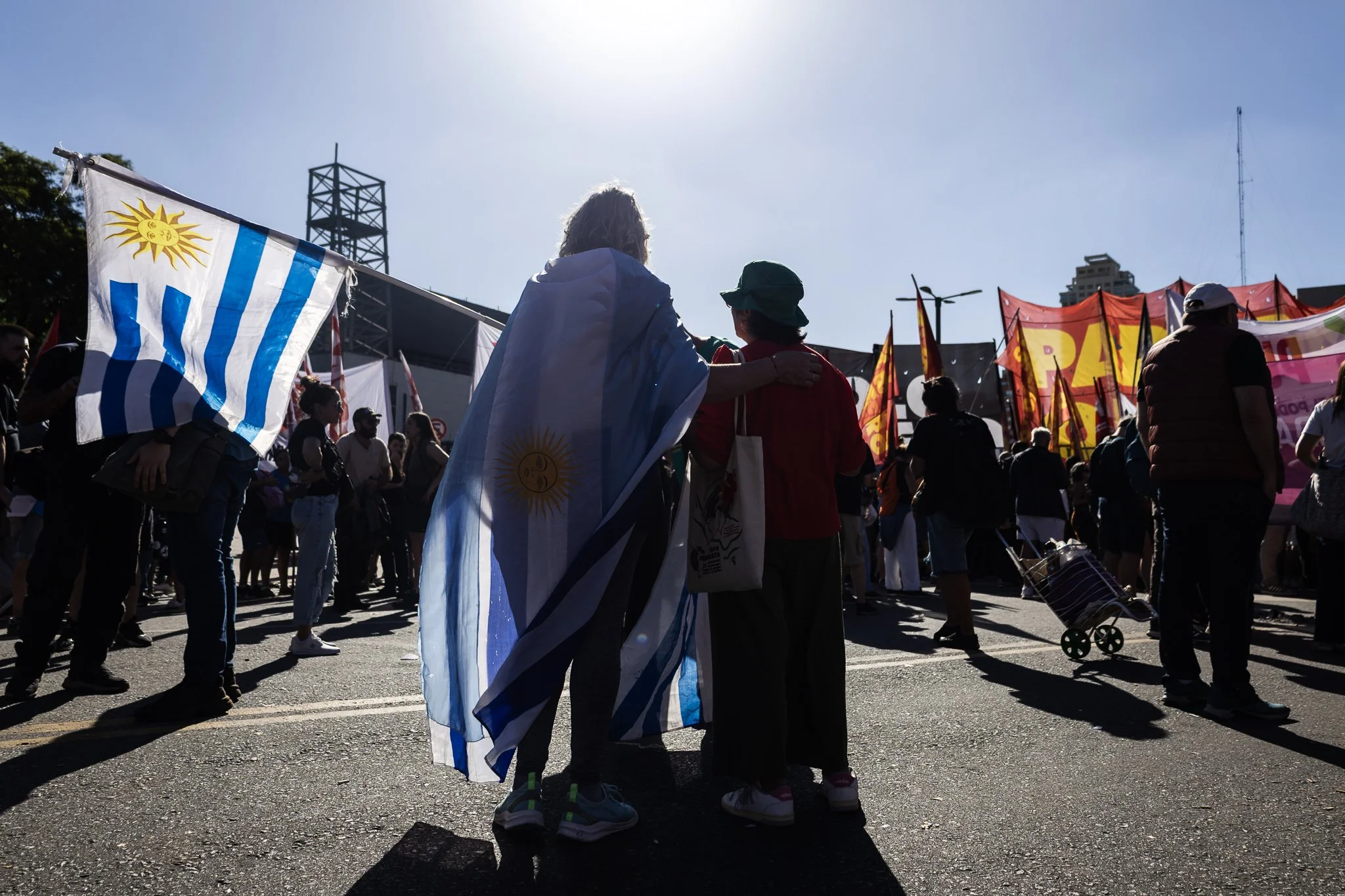 Dos personas en un mitin sosteniendo la bandera de Uruguay, rodeadas de otras personas y banderas en un espacio al aire libre bajo el sol.