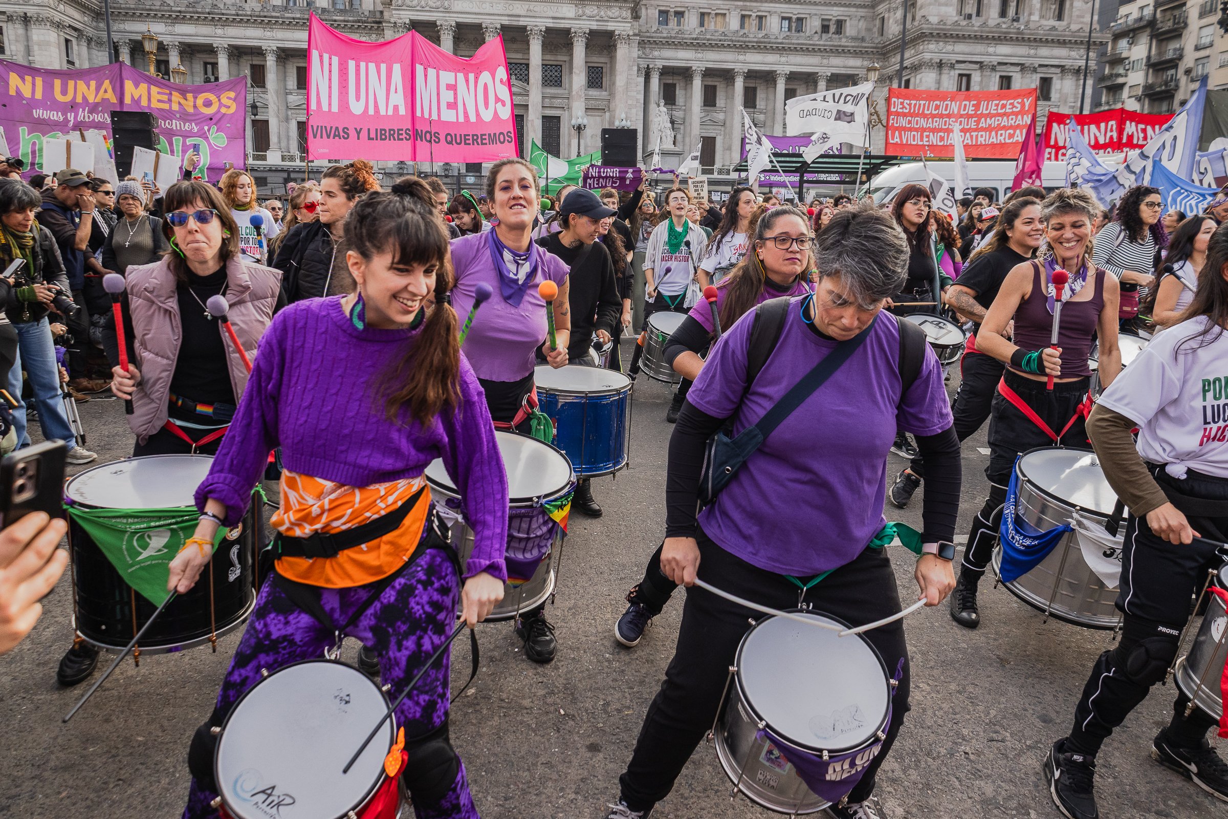 Grupo de mujeres participando en una marcha o manifestación en una plaza, con pancartas que dicen "Ni una menos" y otras frases en contra de la violencia y por los derechos femeninos, con edificios antiguos de fondo.