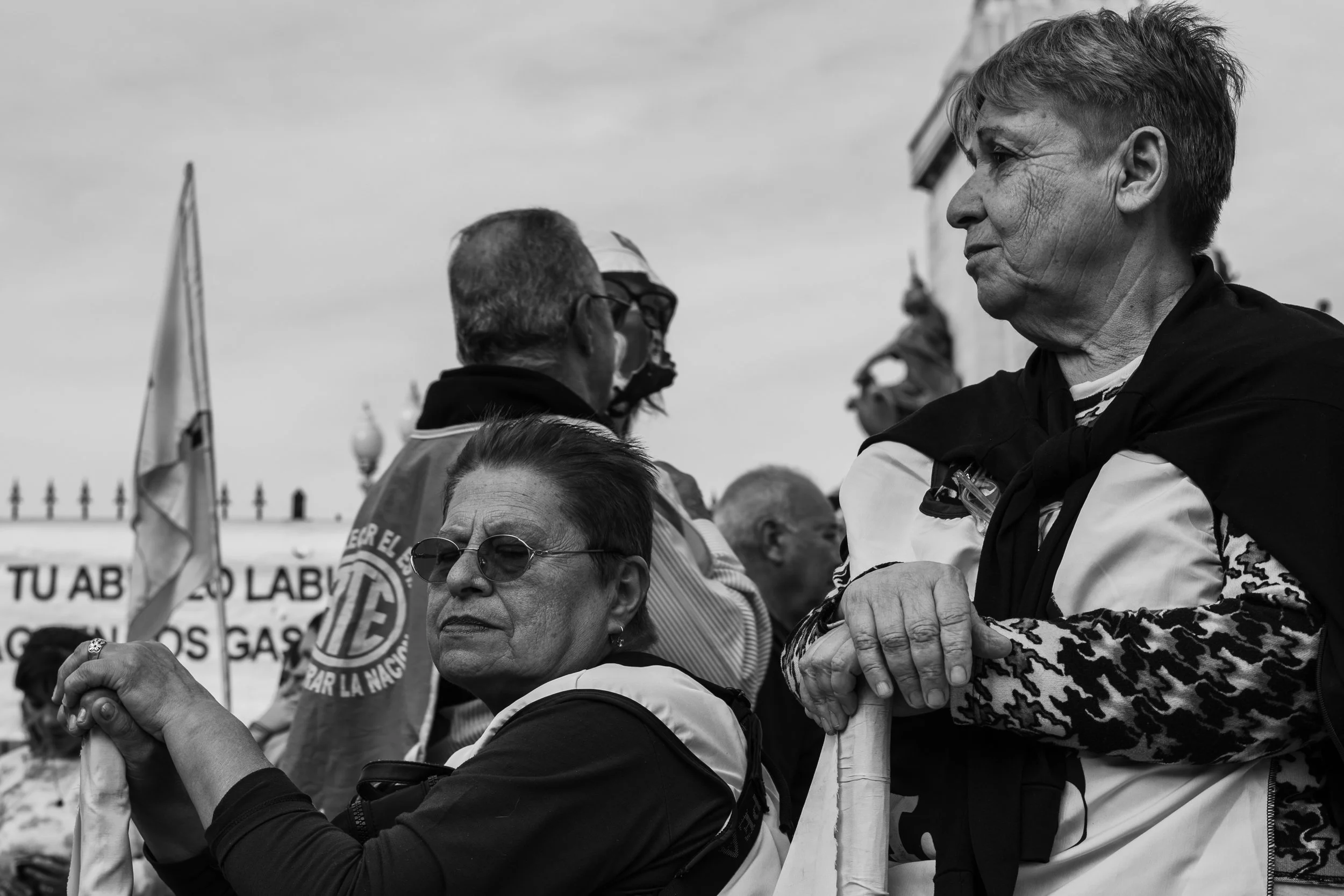 Personas en una manifestación o reunión al aire libre, algunos con pancartas o banderas, en un entorno urbano con vallas y un cielo nublado.