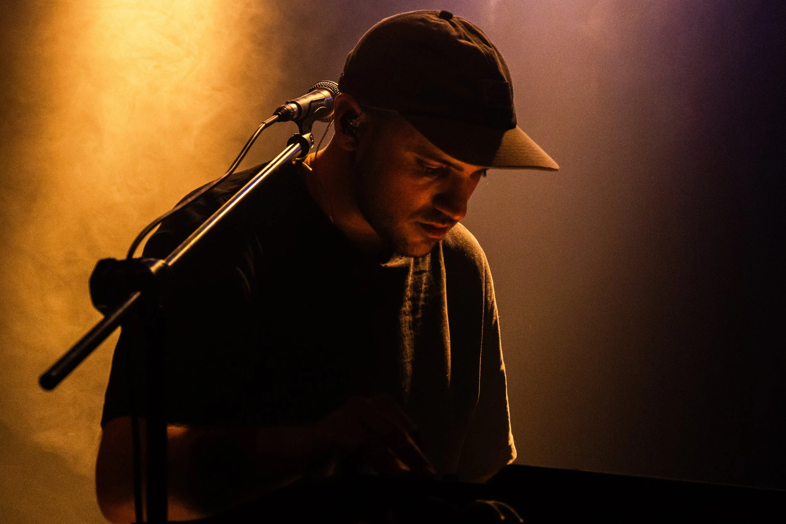 Hombre con gorra oscuro en un escenario con iluminación cálida, de espaldas a un micrófono, en un concierto .