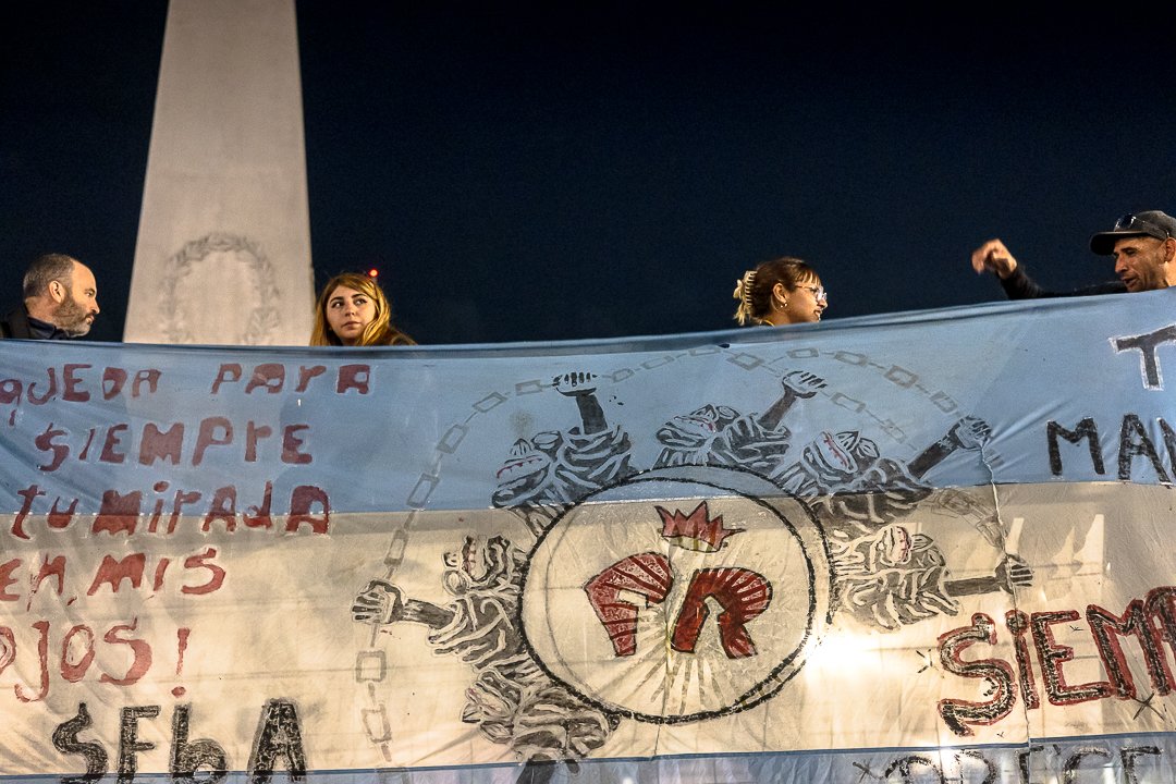 Grupo de personas sosteniendo una pancarta con un símbolo de grafiti que incluye una corona y letras, en un ambiente nocturno.