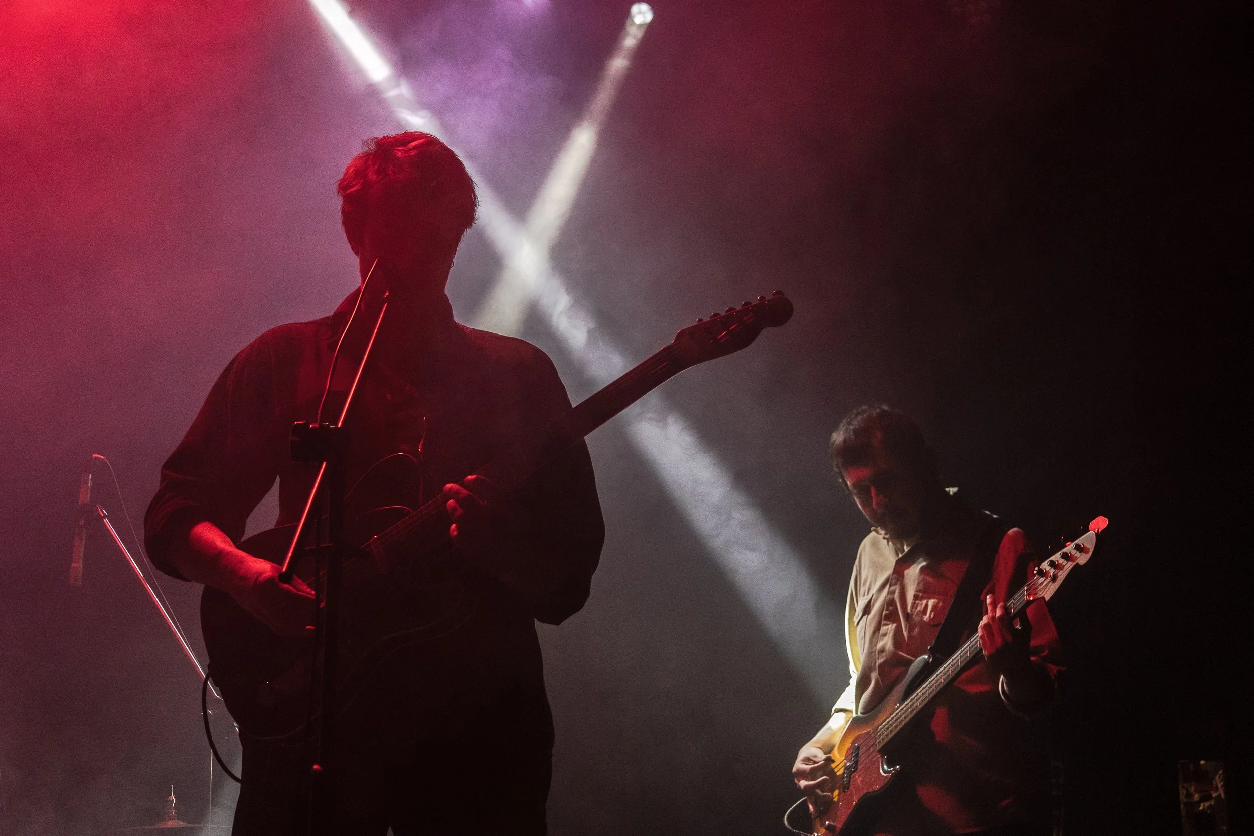 Dos músicos tocando en un escenario con luces de fondo, uno con guitarra eléctrica y otro con guitarra acústica, en un ambiente de concierto en vivo.