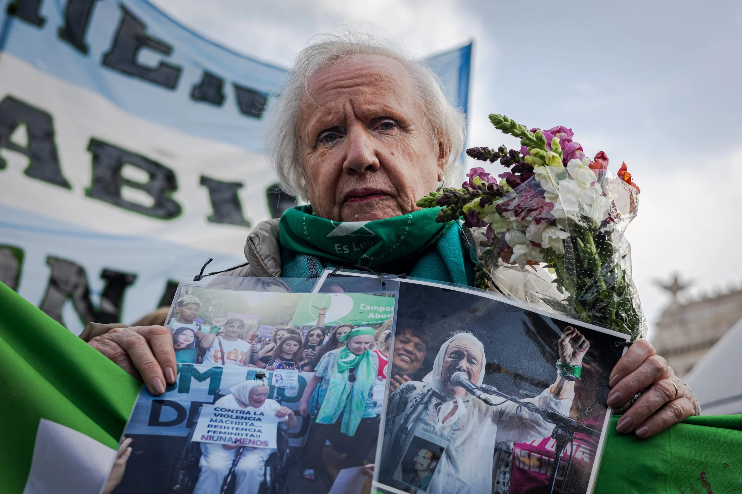 Una mujer mayor en una protesta, sosteniendo fotos y un cartel que protesta contra la violencia machista, lleva un pañuelo verde y una flor en la mano.