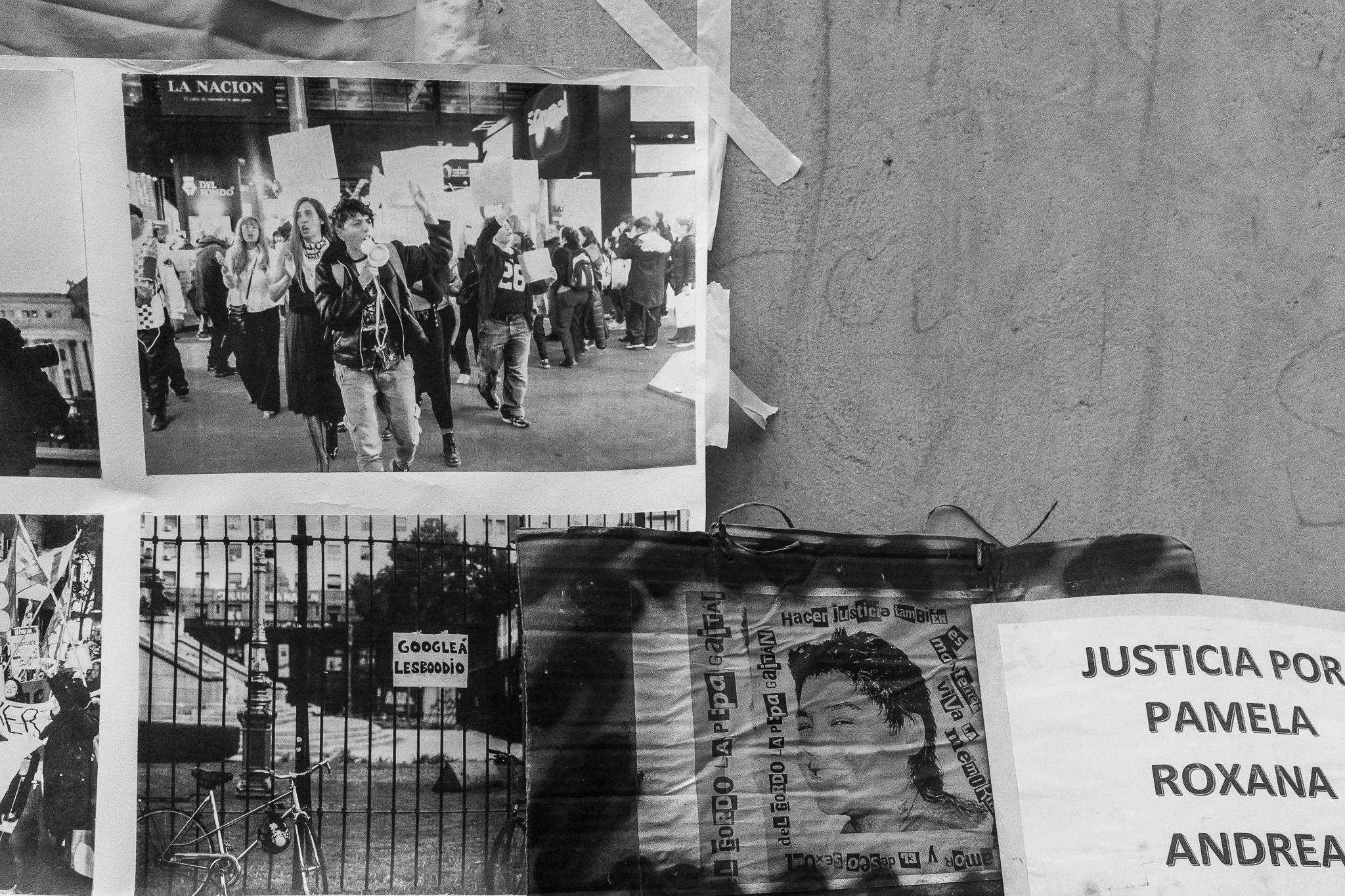 Fotografías en blanco y negro en una pared de manifestaciónes con varias personas caminando, algunas con carteles. Al lado, un cartel dice ''Justicia por Pamela, Roxana y Andrea'.