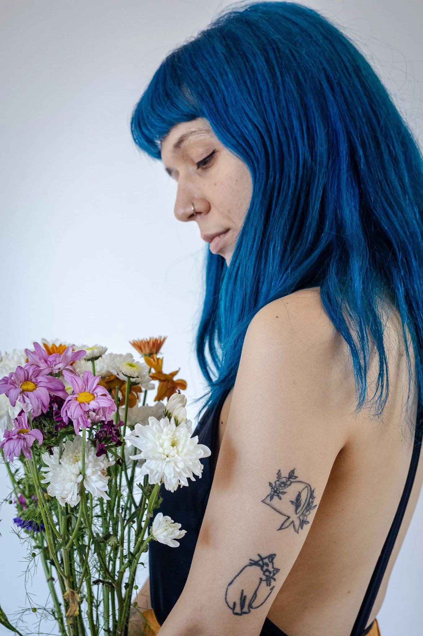 Mujer con cabello azul sosteniendo un ramo de flores coloridas, tiene tatuajes en el brazo y lleva un vestido negro con espalda descubierta.