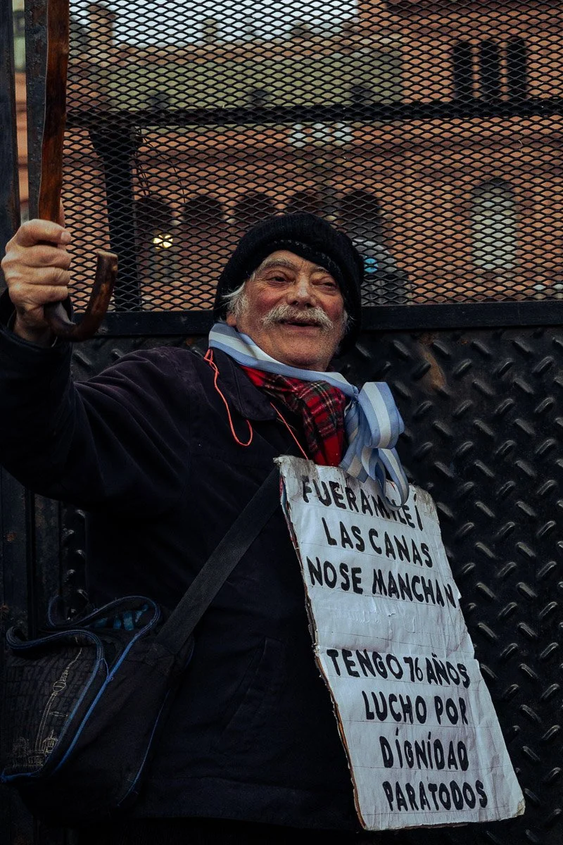 Un hombre mayor con barba y sombrero negro mostrando una sonrisa, lleva un cartel colgado que dice 'Fuera Merkel, las canas no se manchan, tengo 76 años, lucho por la dignidad para todos'.