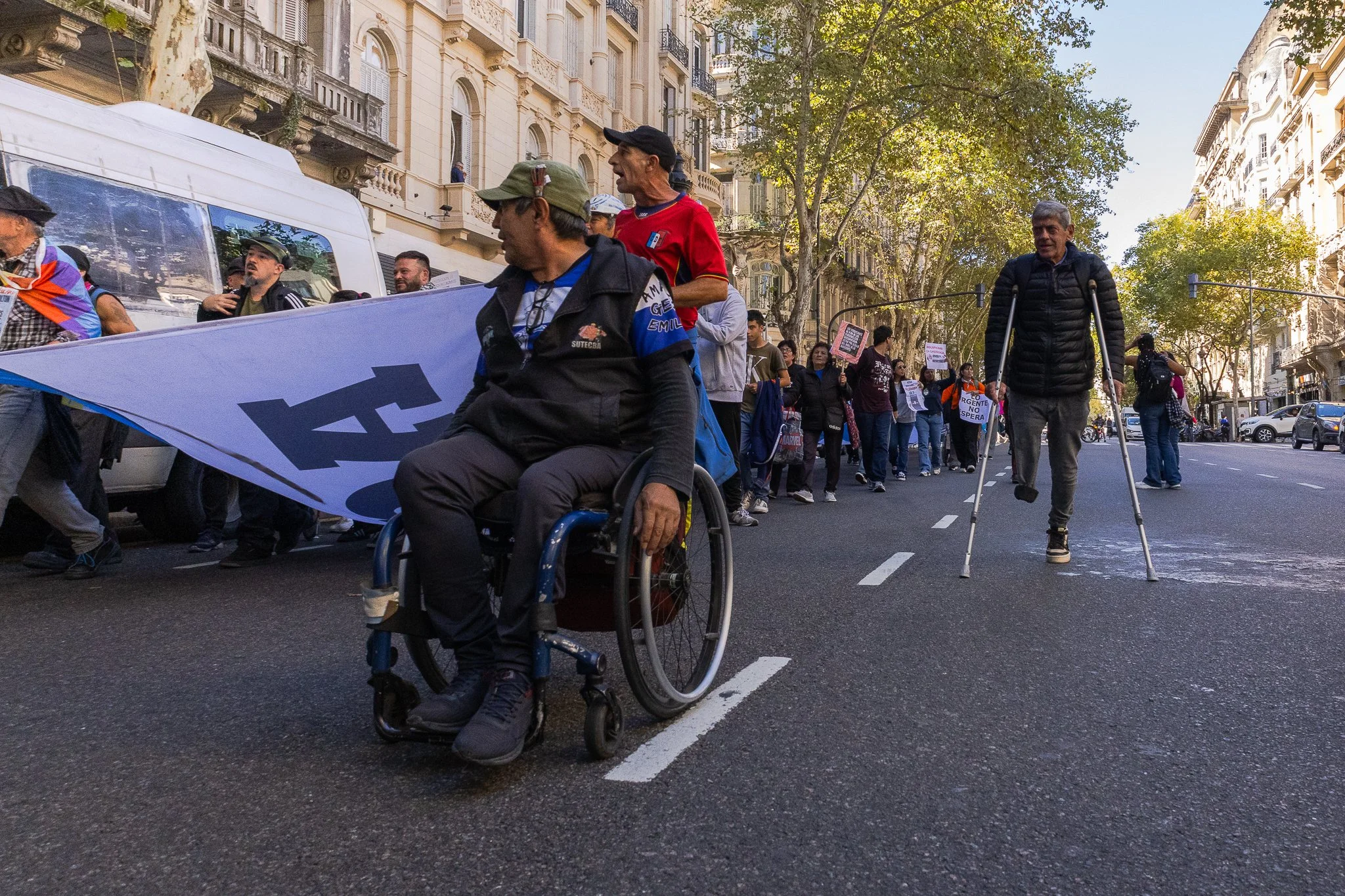 Grupo de personas participando en una marcha o manifestación en una calle urbana, algunas llevando pancartas y banderas, con árboles y edificios antiguos en el fondo.