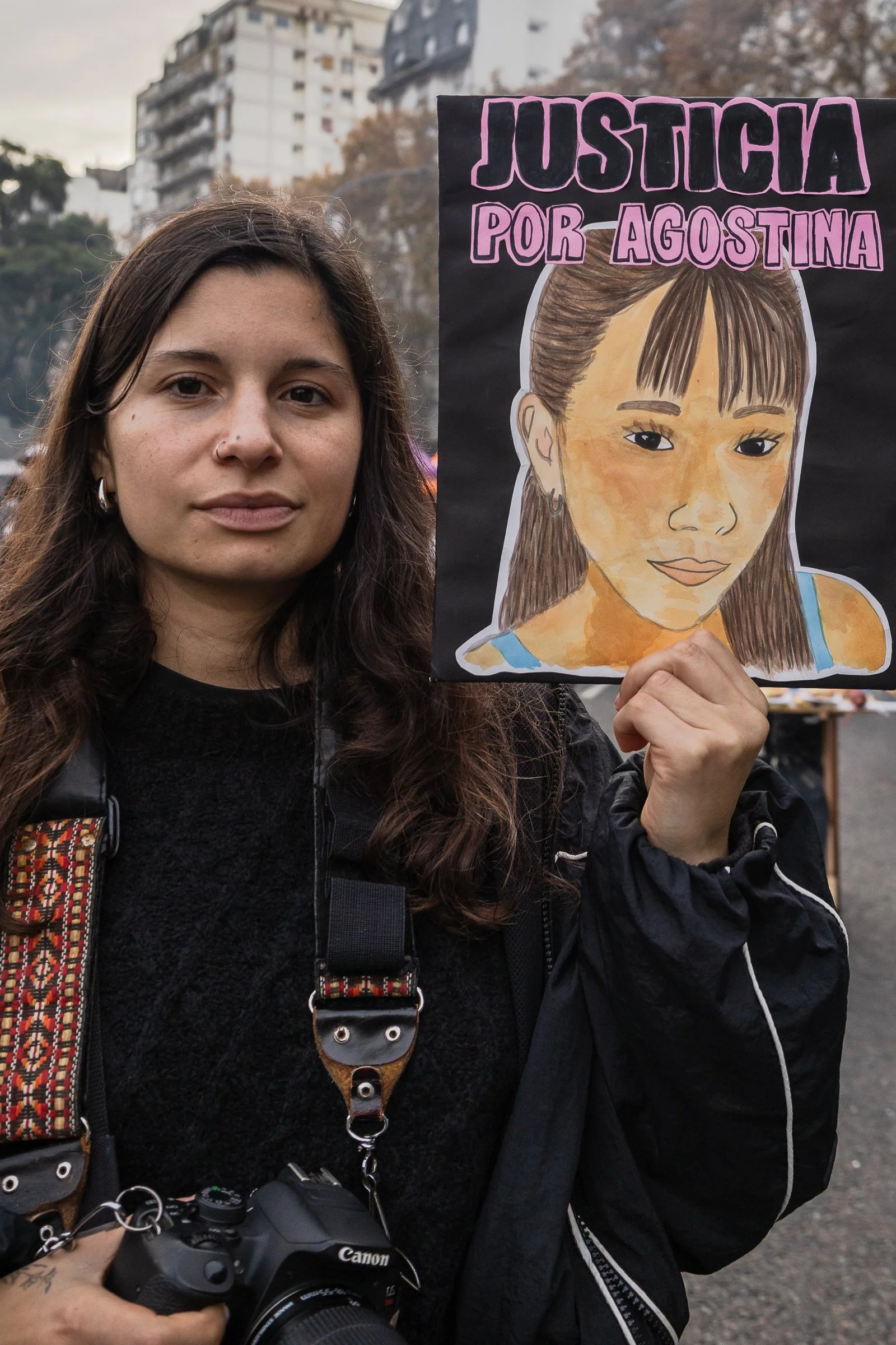 Mujer con cabello largo oscuro y pava en un árbol, sosteniendo cartel con retrato de niña y frase 'Justicia por Agostina' en una manifestación en exteriores.