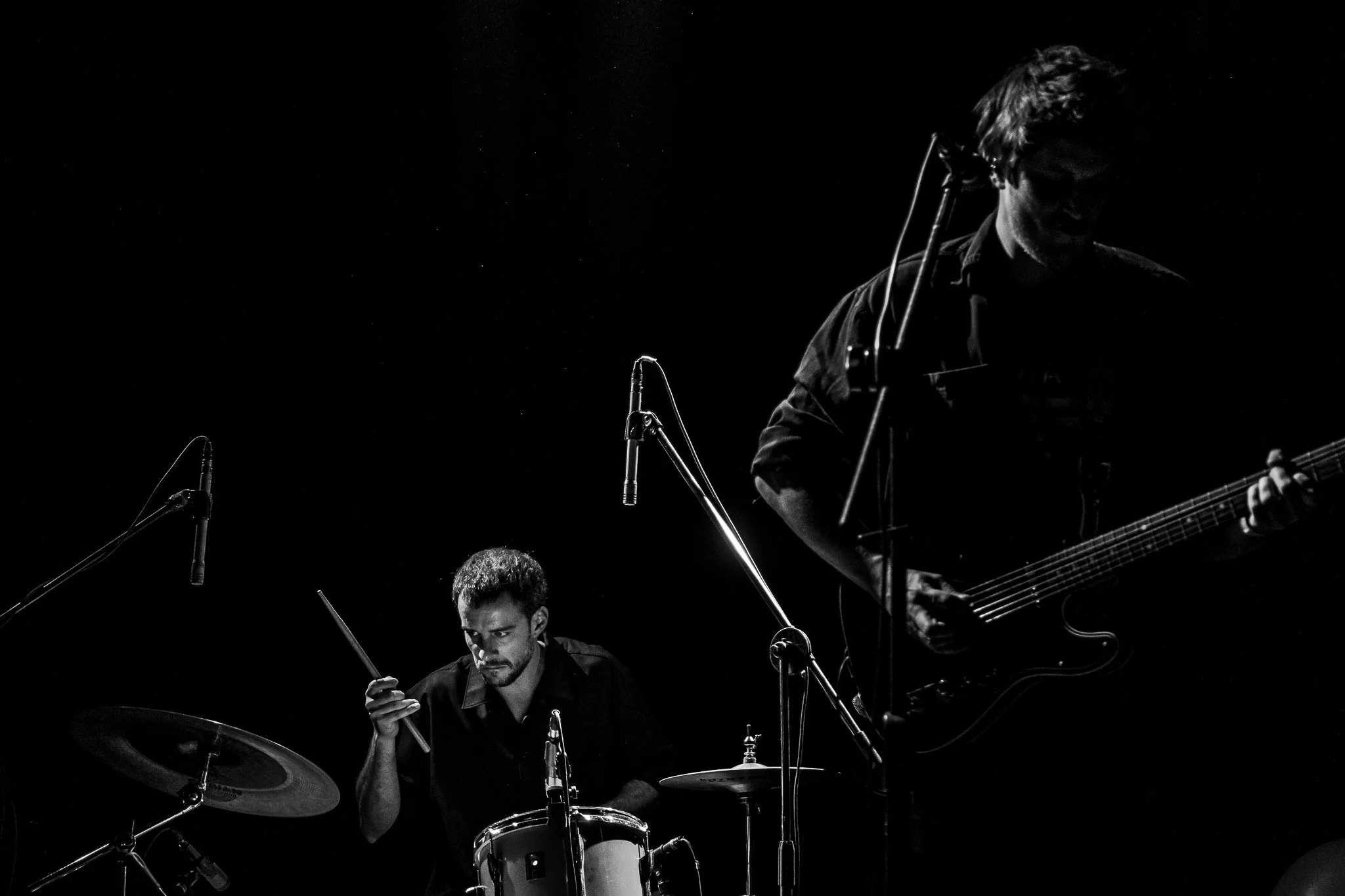 Músicos tocando instrumentos en un escenario oscuro, uno con guitarra y otro con batería.