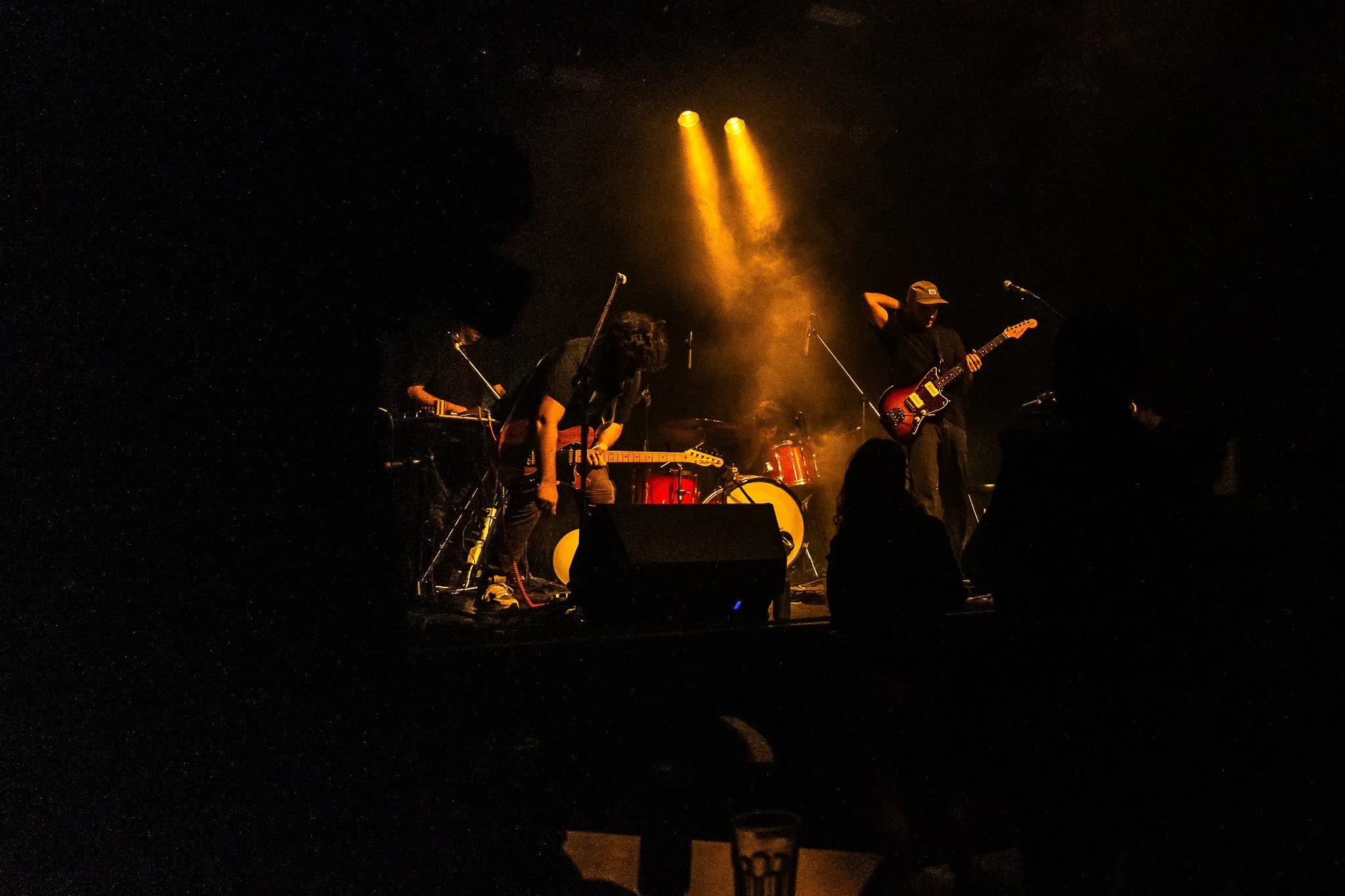 Un grupo de músicos tocando en un escenario oscuro con iluminación amarilla, tocando guitarras y batería.