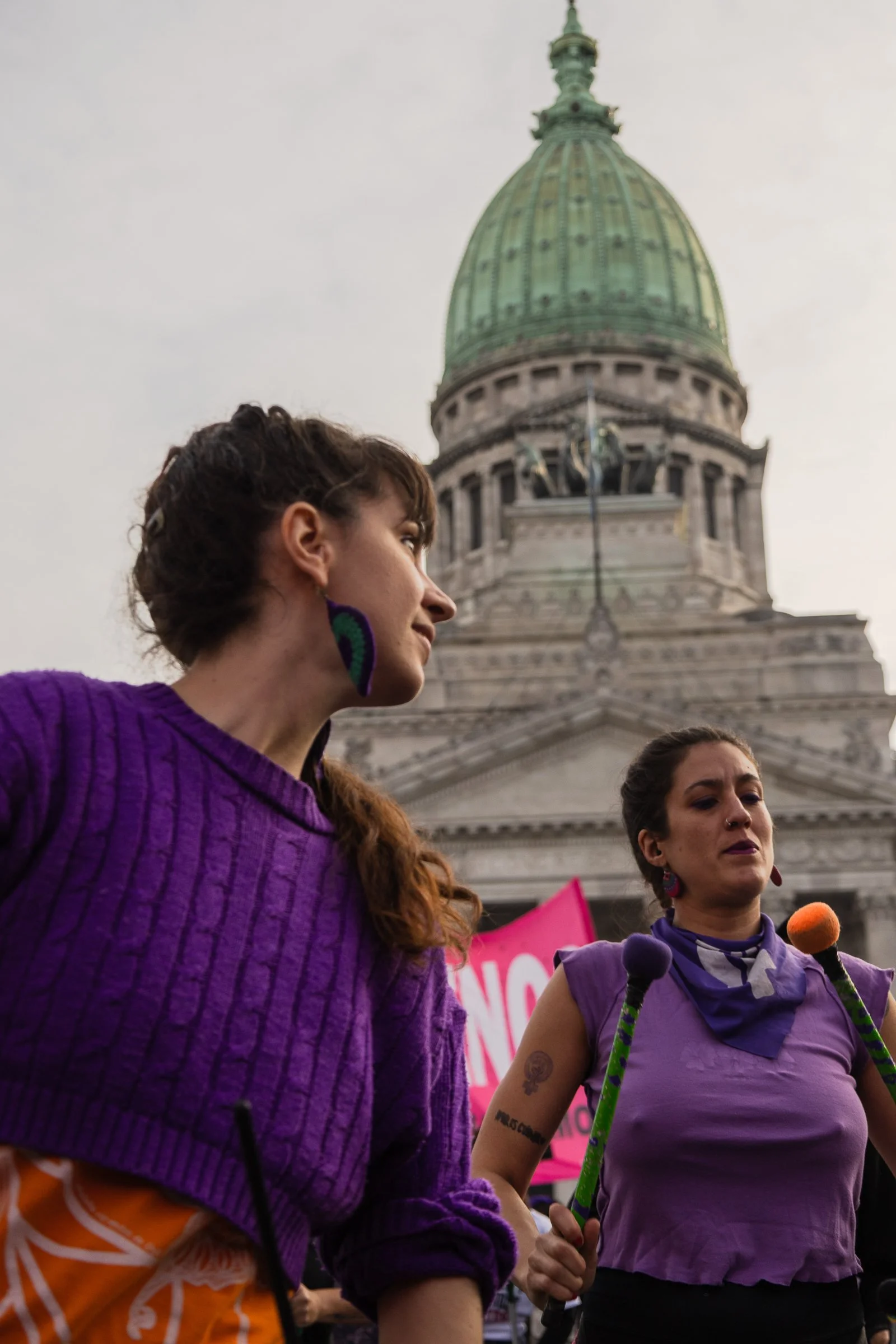 Mujeres participando en una manifestación frente a un edificio con cúpula grande, vestidas con ropa morada, en un ambiente de protesta.