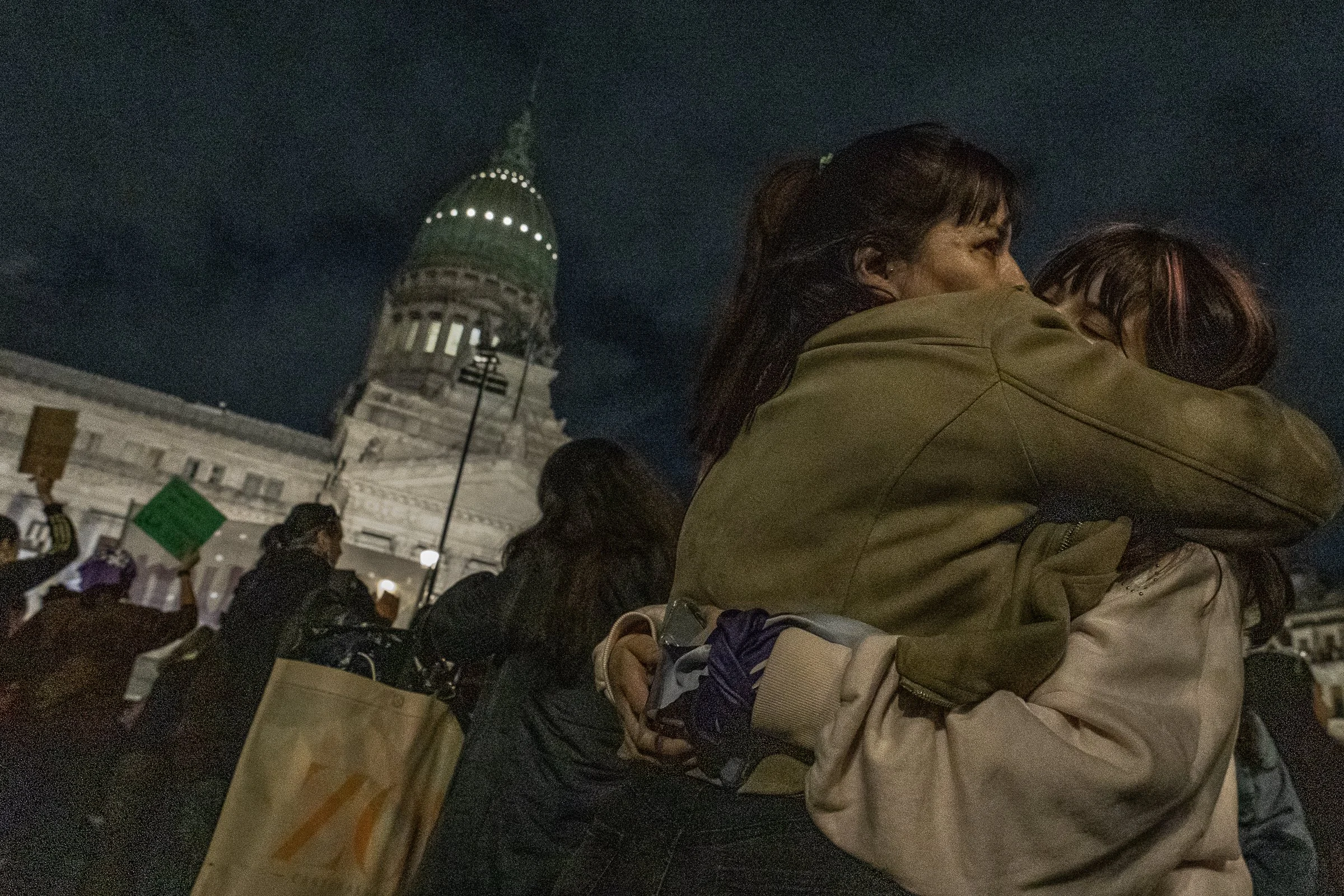 Madre e hija abrazándose en una manifestación nocturna frente a un edificio con cúpula dorada.