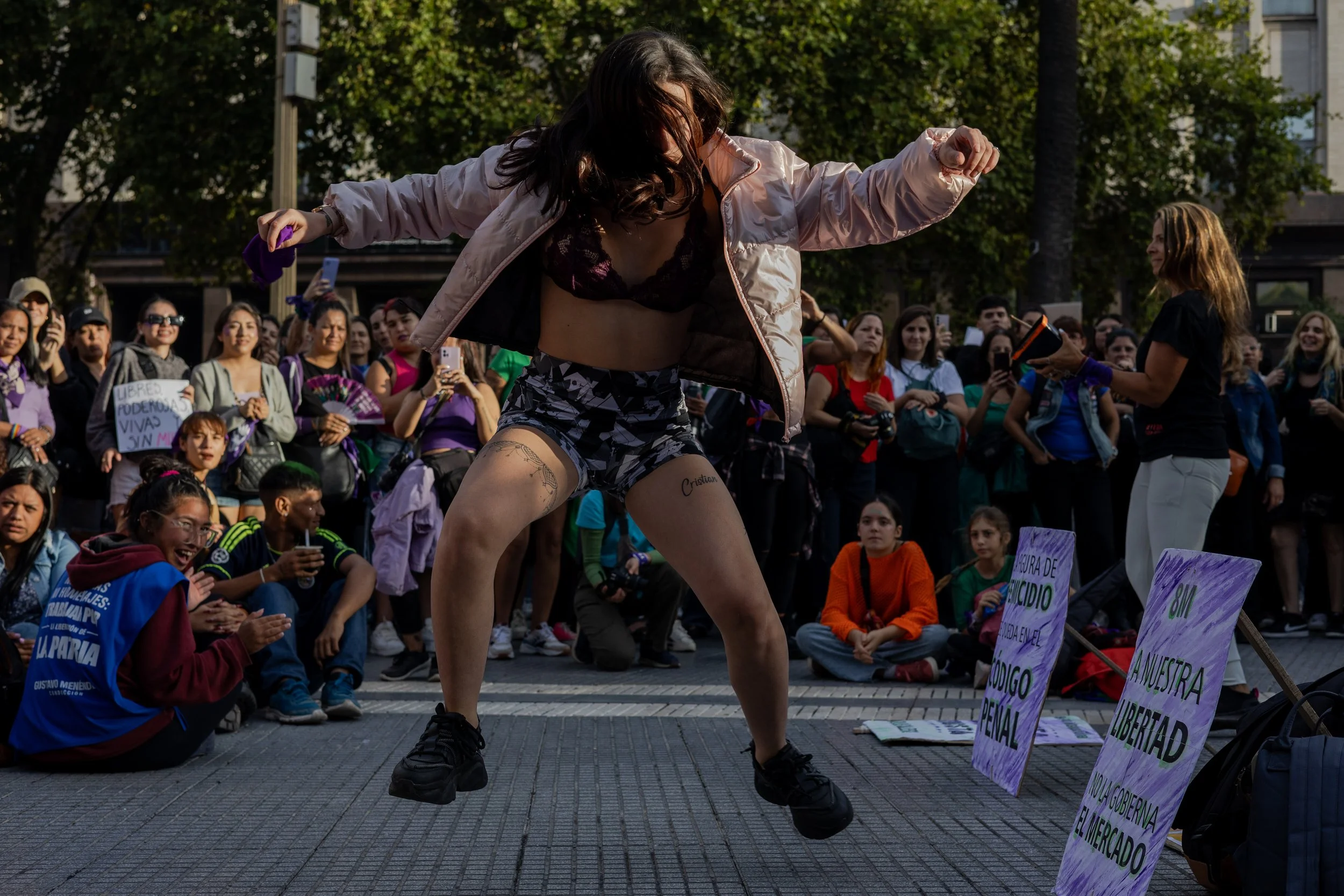 Una mujer bailando en una protesta o evento público rodeada de un público que la observa, algunos tomándose fotos, en un espacio al aire libre con árboles y edificios al fondo.