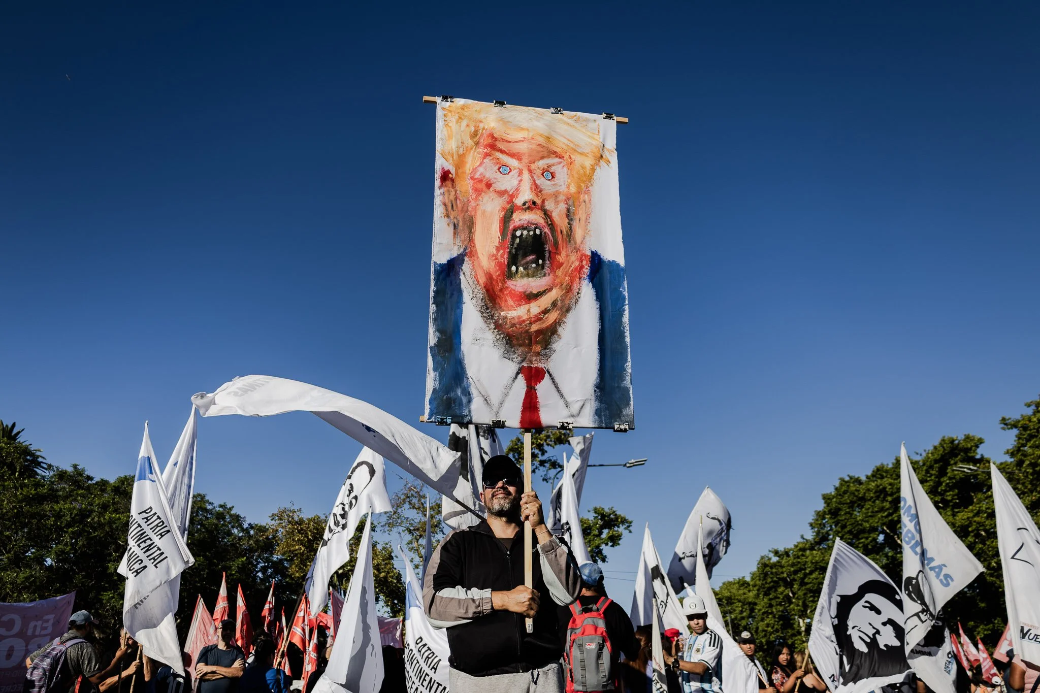Persona sosteniendo una pancarta con un dibujo caricaturesco de un hombre con cabello rubio y expresión de enojo en un mitin con banderas y personas en el fondo.