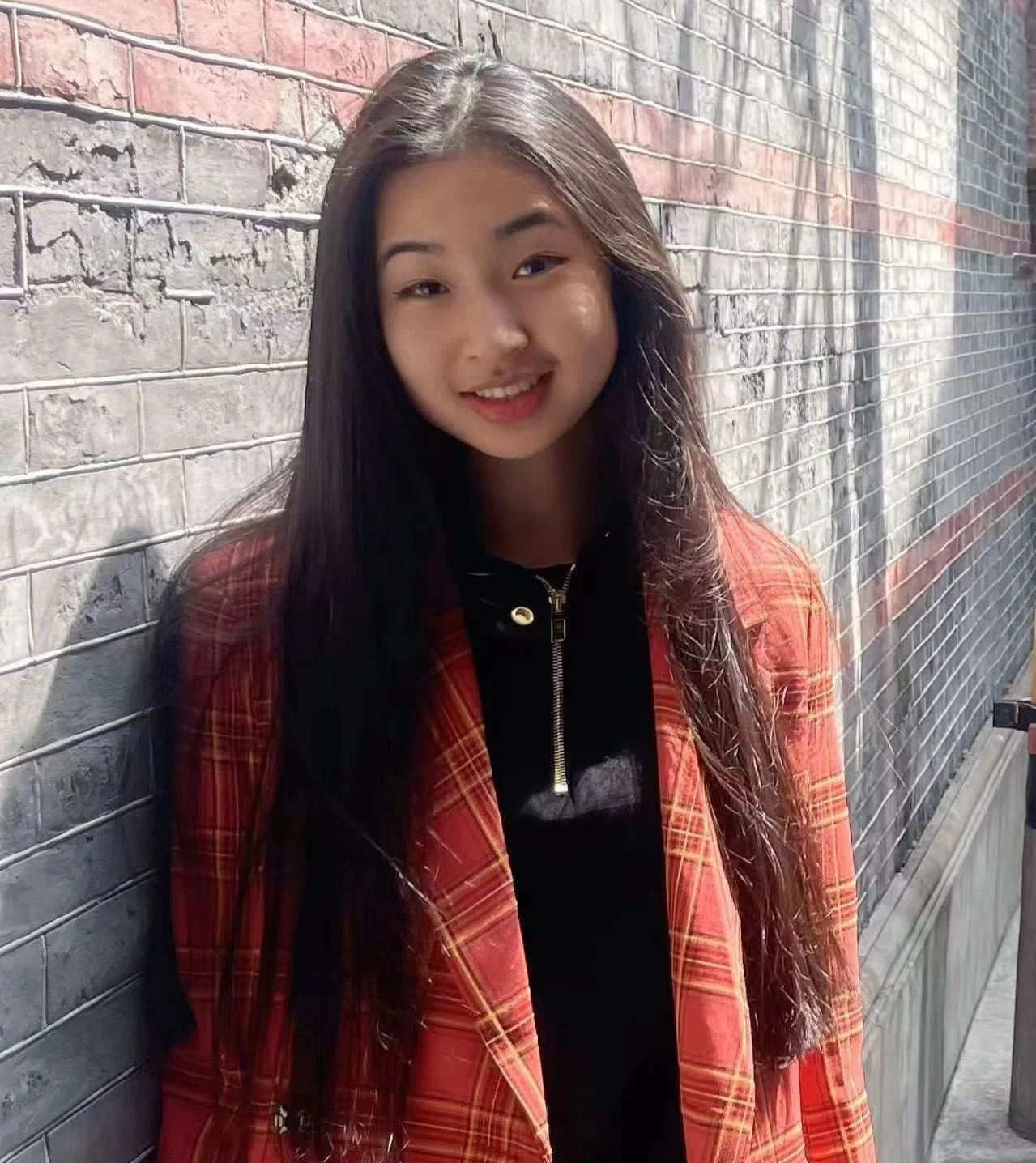 A young woman with wavy brown hair, is smiling and standing outdoors near green plants.