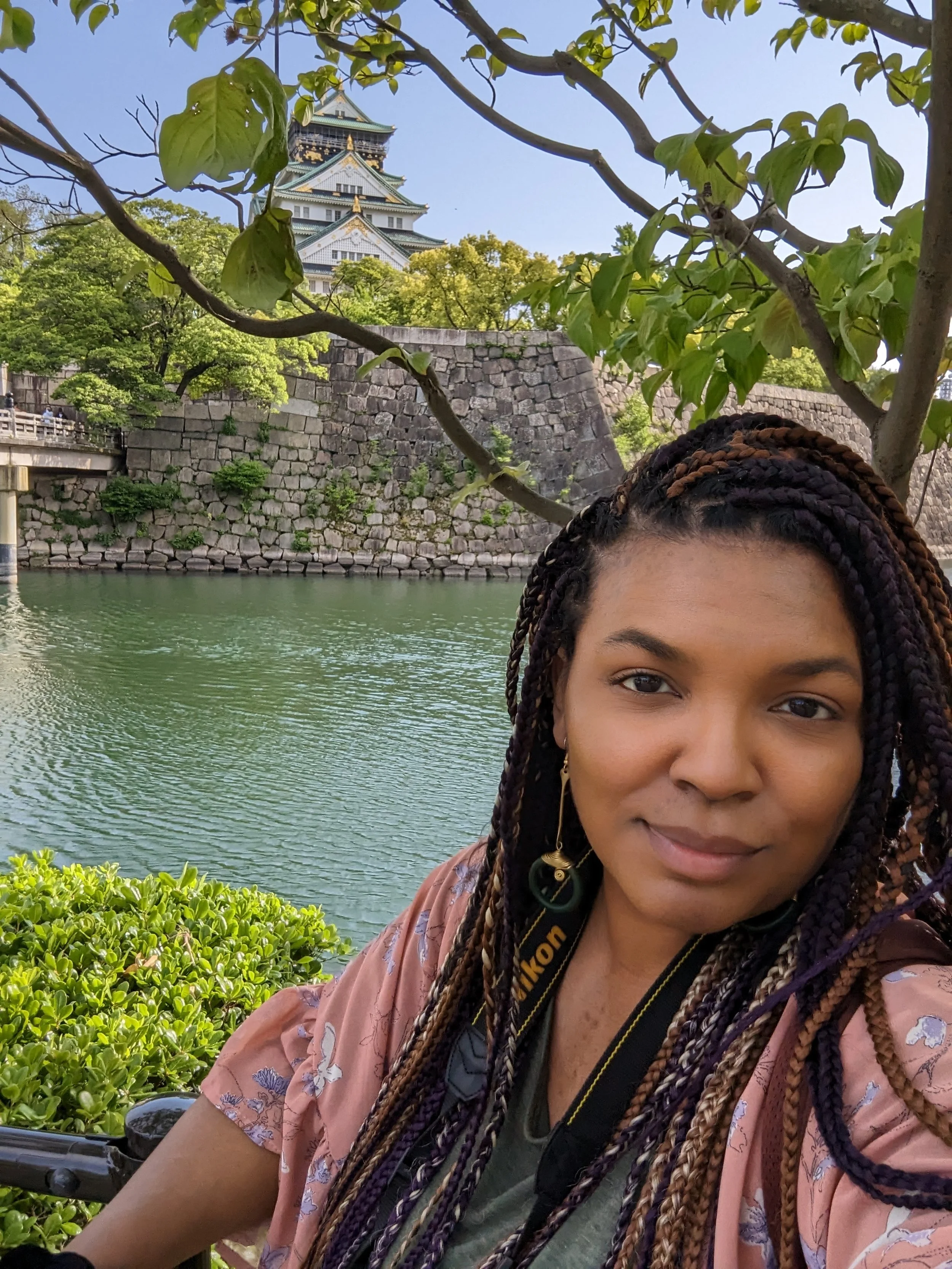 A  Black woman with box braids in her hair taking a selfie outdoors near a body of water with a stone wall and traditional Japanese castle in the background.