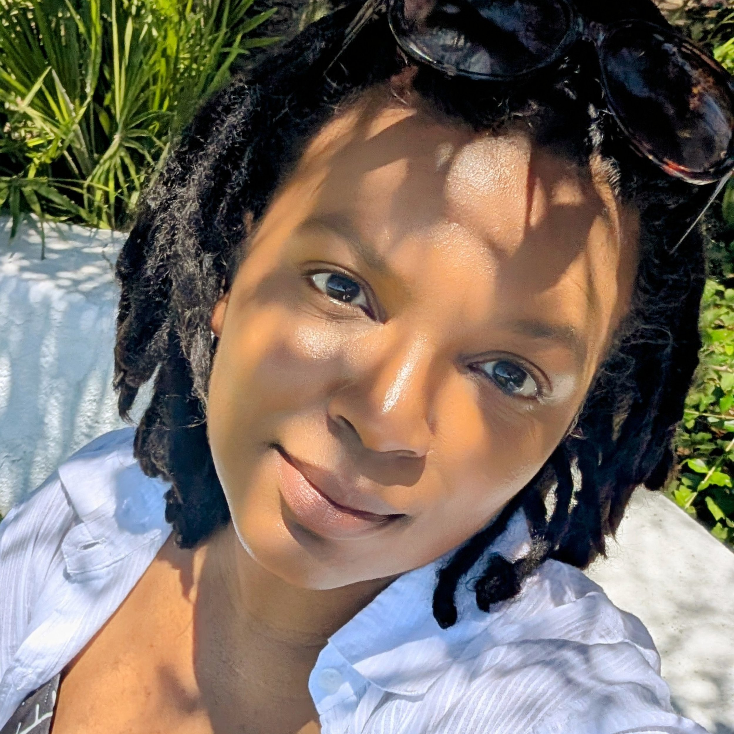 A Black woman takes a selfie outdoors in sunlight, wearing sunglasses on her head and a white shirt, with green plants in the background.