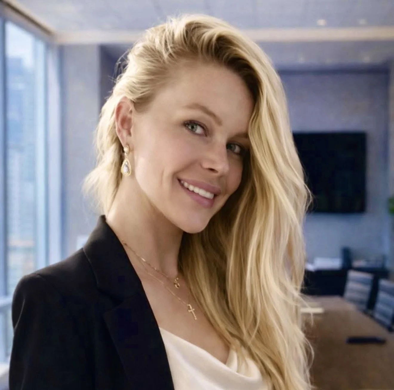 Allison Bonham smiling blonde woman with wavy hair, wearing earrings, a necklace with a cross pendant, a black blazer, and a white top, standing in a modern office with large windows and a city view.