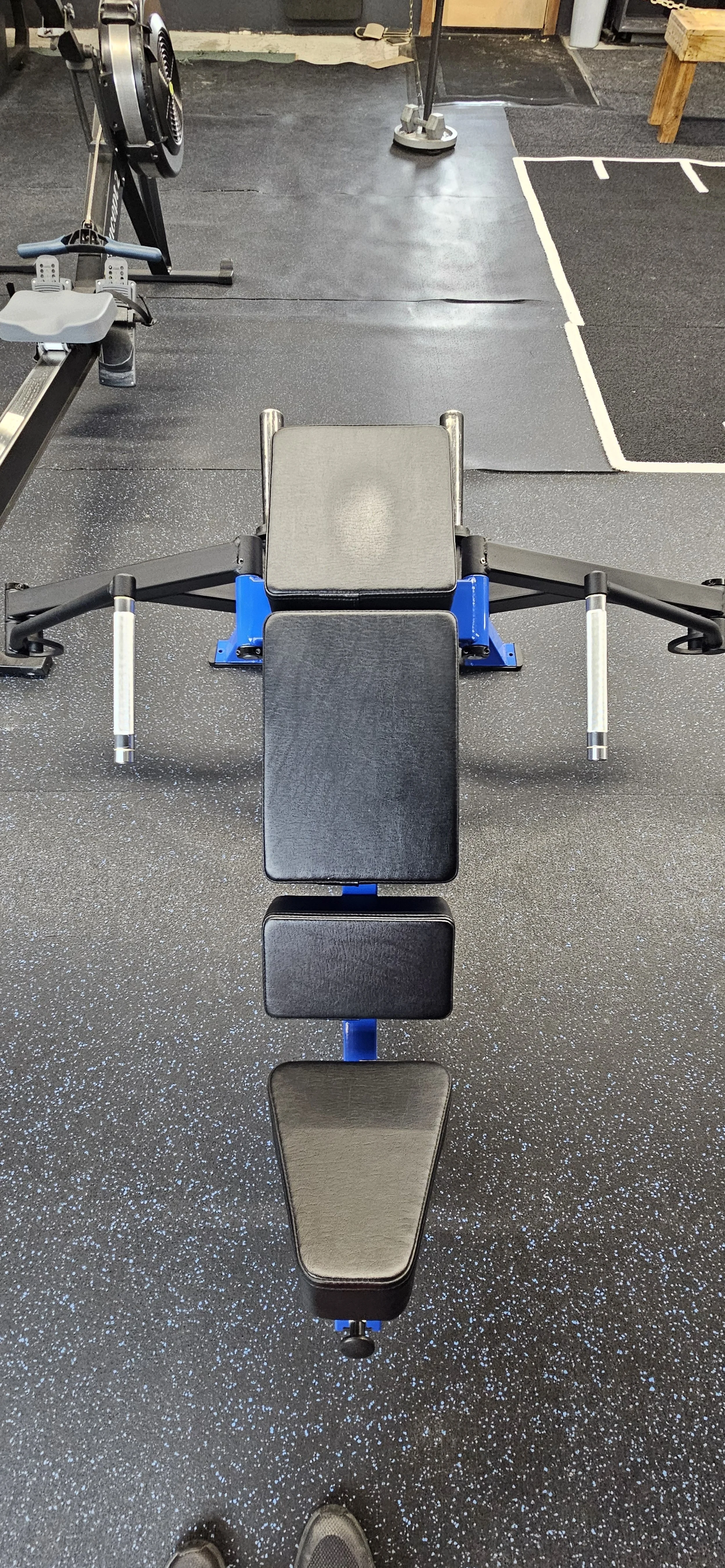 A black adjustable workout bench with padded sections in a gym. The gym has black rubber flooring with small white specks and various exercise equipment in the background.