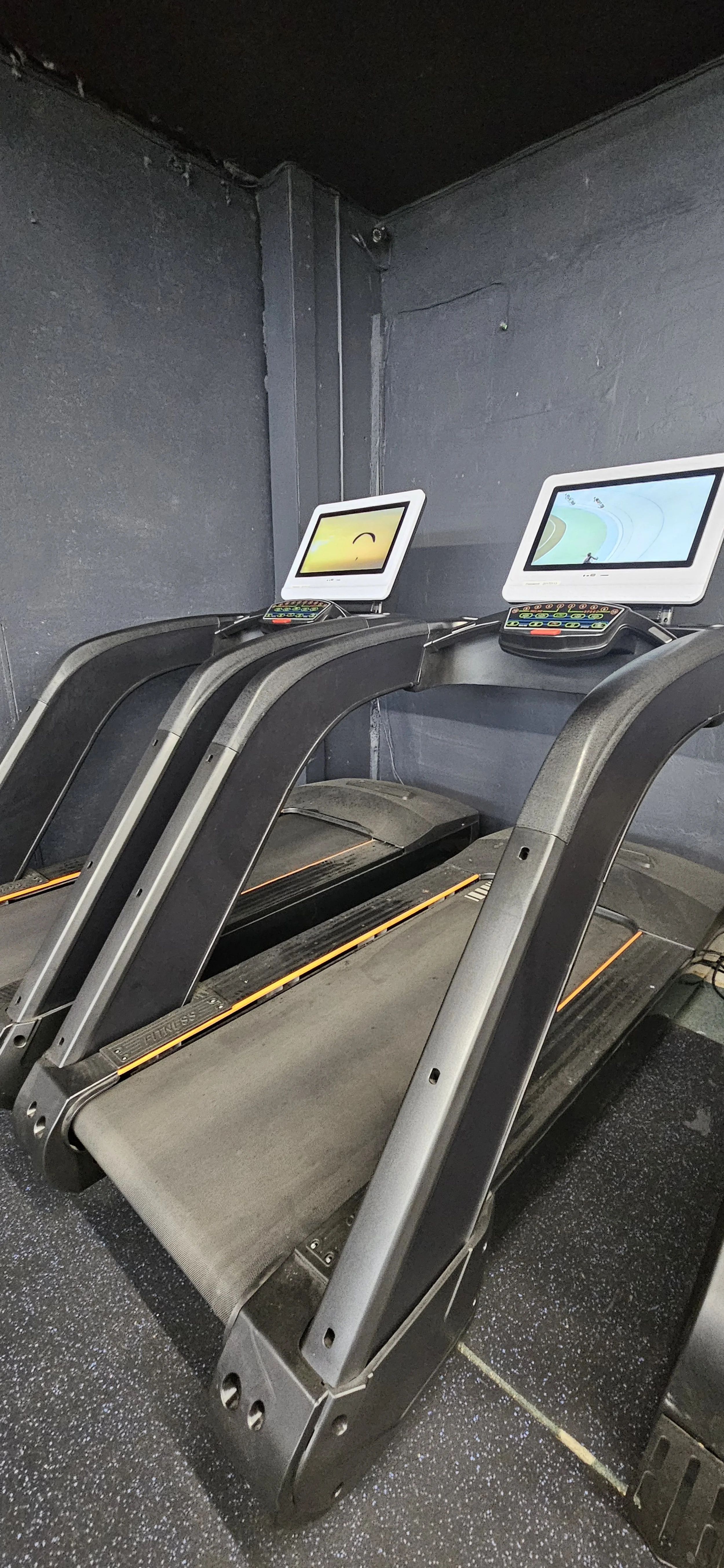 Three black and gray treadmills with digital screens in a dark gym corner with black wall and floor.