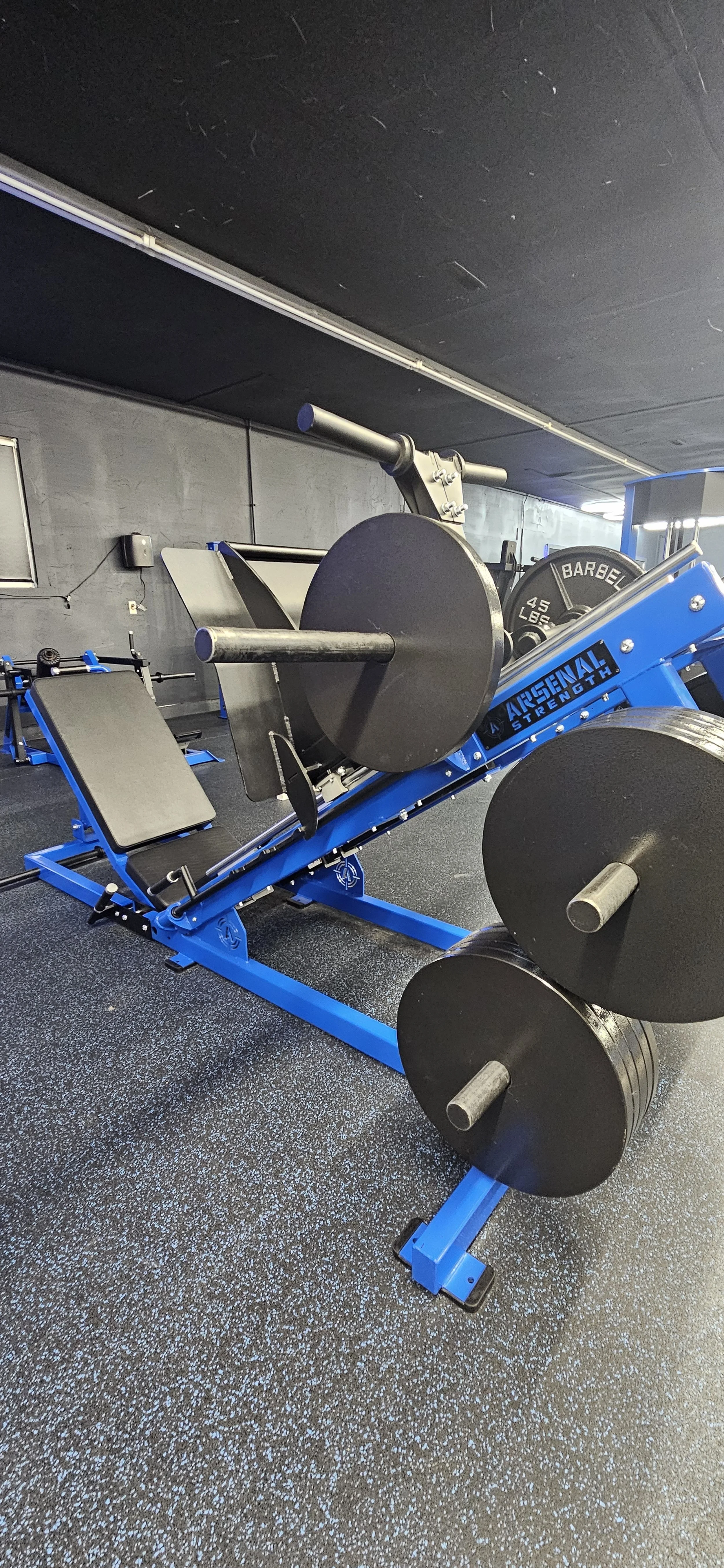 A blue Argential Strength workout machine in a gym with black weights and a black wall.